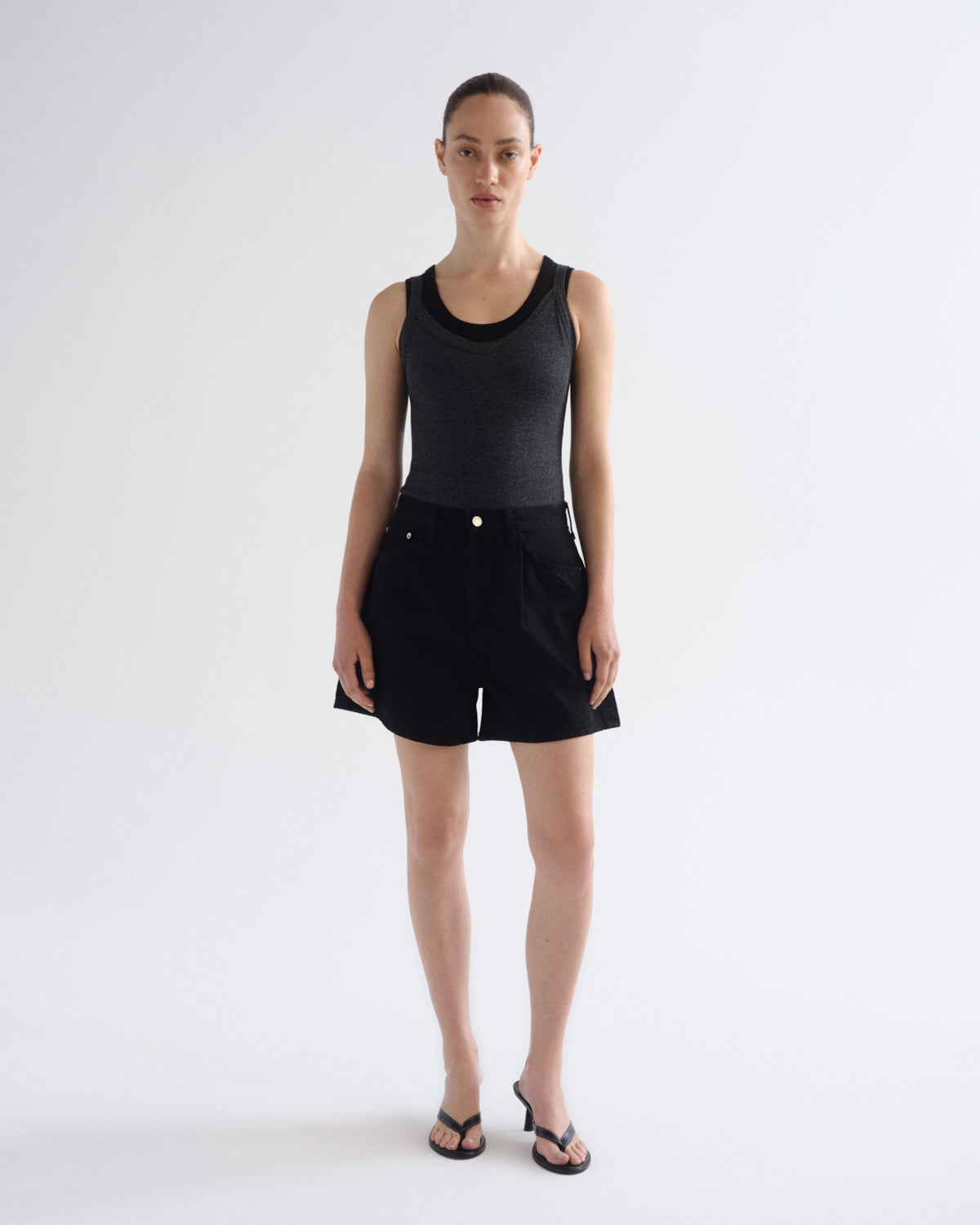 A model stands against a plain light gray studio background wearing a black ribbed tank top and black shorts, with black thong sandals.