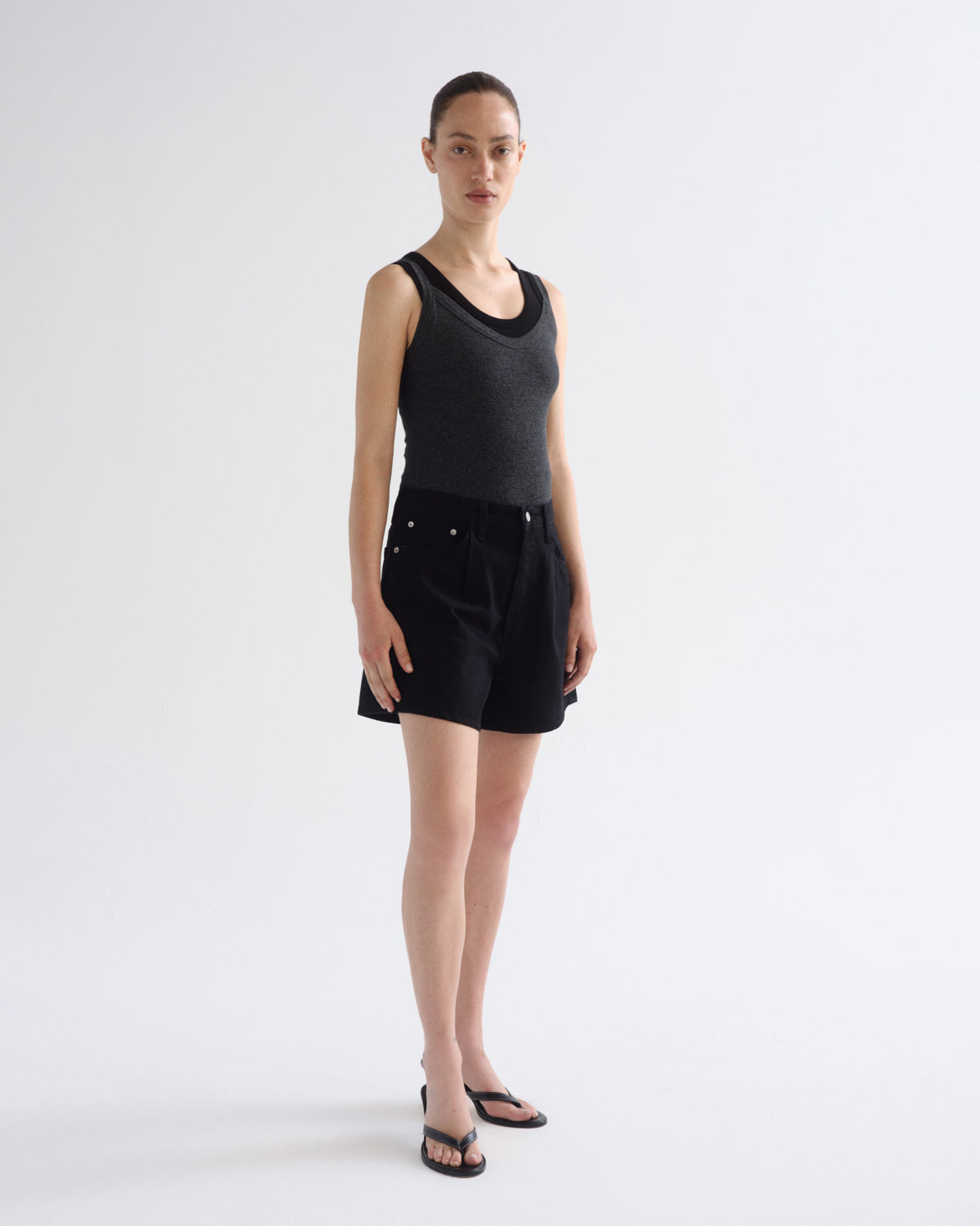 A model stands against a light gray studio background wearing a dark charcoal gray ribbed tank top and black shorts, paired with black sandals.