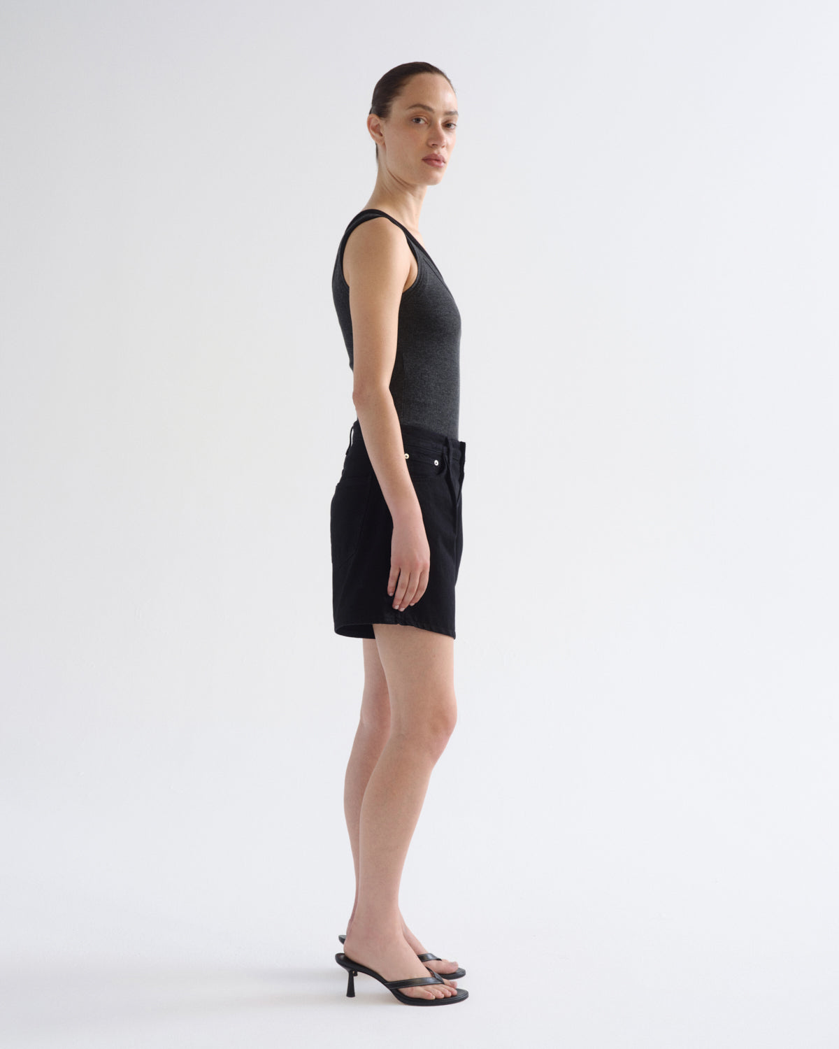 A woman stands in profile wearing a black sleeveless top and black shorts against a clean white studio background.