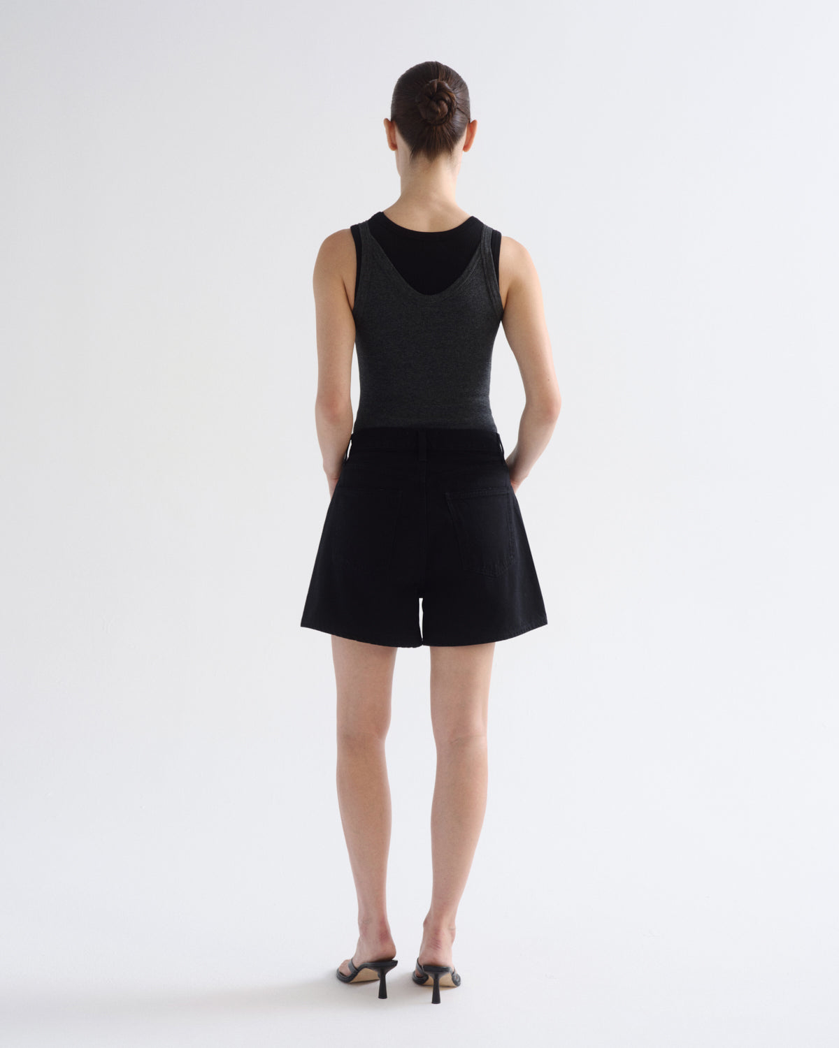 A model shown from the back wearing a sleeveless black top with a textured back panel and black shorts, paired with black heeled sandals, against a plain white studio background.