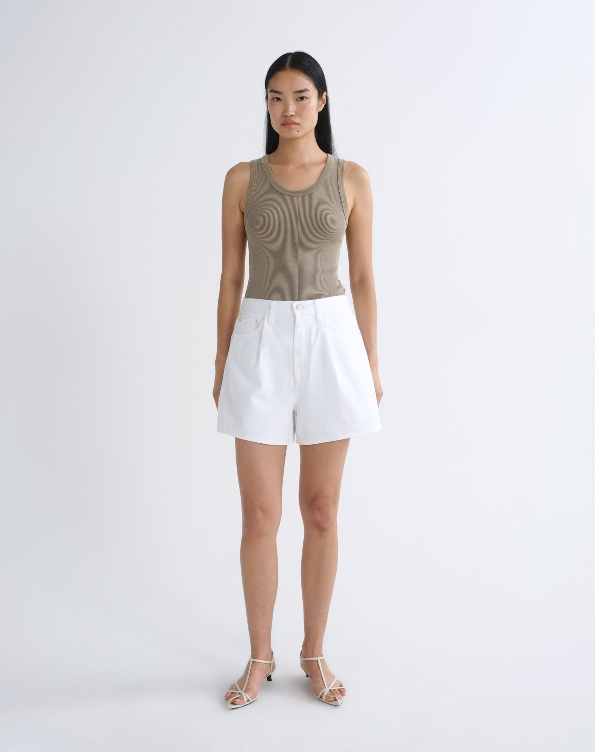 A model stands against a light gray studio background wearing a sleeveless taupe-beige ribbed tank top, white shorts, and silver strappy heels.