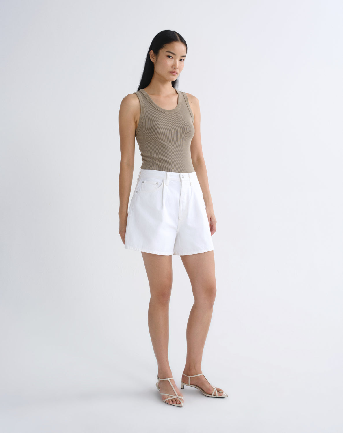 A model stands against a light gray studio background wearing a beige ribbed tank top, white shorts, and clear-strap heeled sandals.