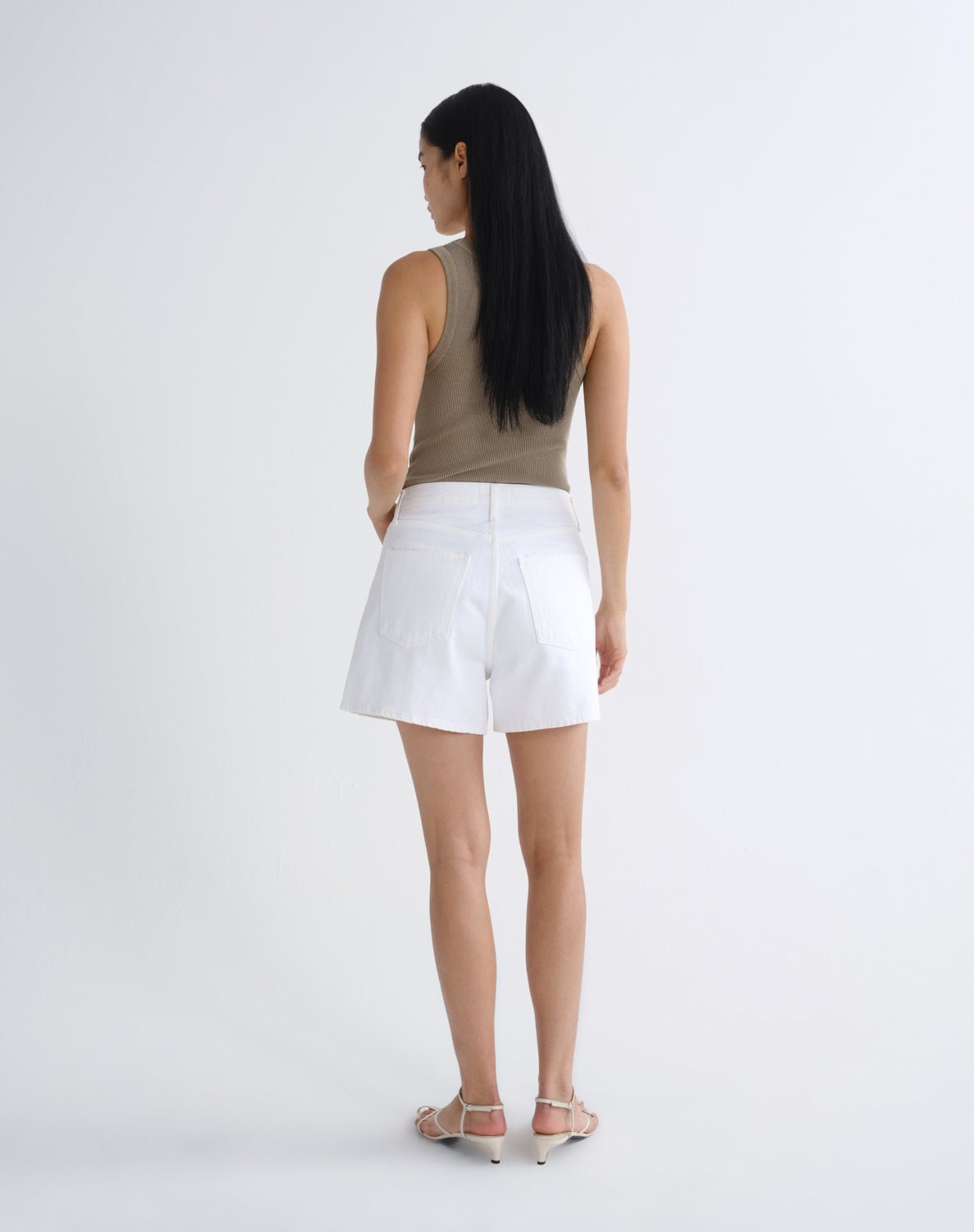 A woman with long black hair is shown from behind wearing white shorts, a taupe sleeveless top, and light-colored high-heeled sandals against a plain white studio background.