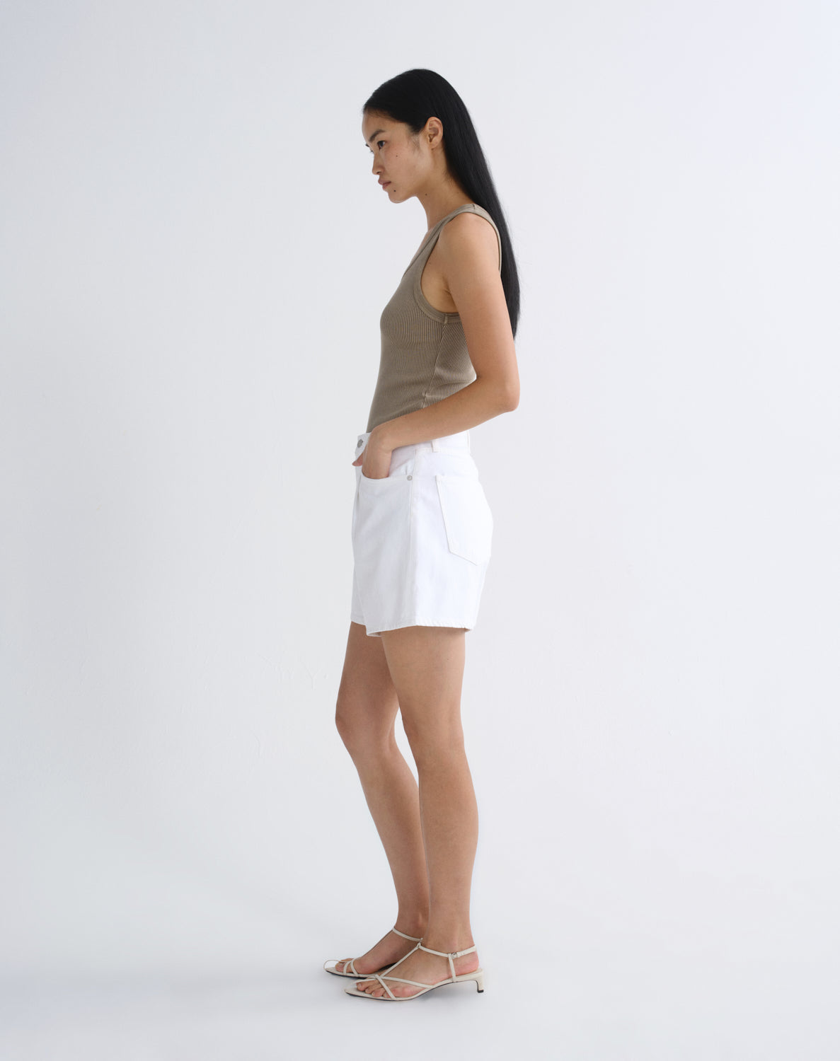 A woman stands in profile wearing a sleeveless taupe top, white shorts, and strappy high-heeled sandals against a clean white studio background.