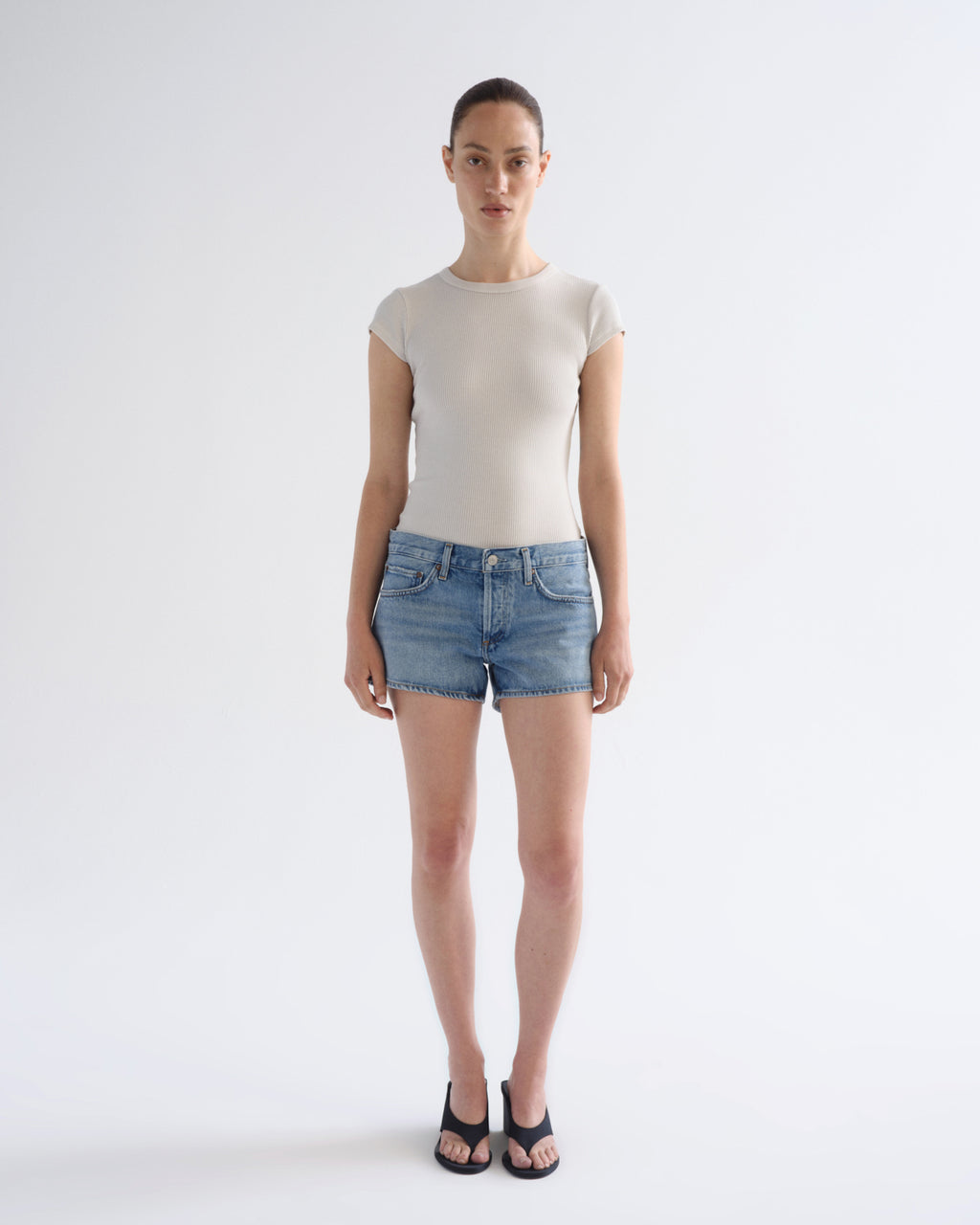 A model stands against a light gray studio background wearing a fitted short-sleeve beige top, blue denim shorts, and black sandals.