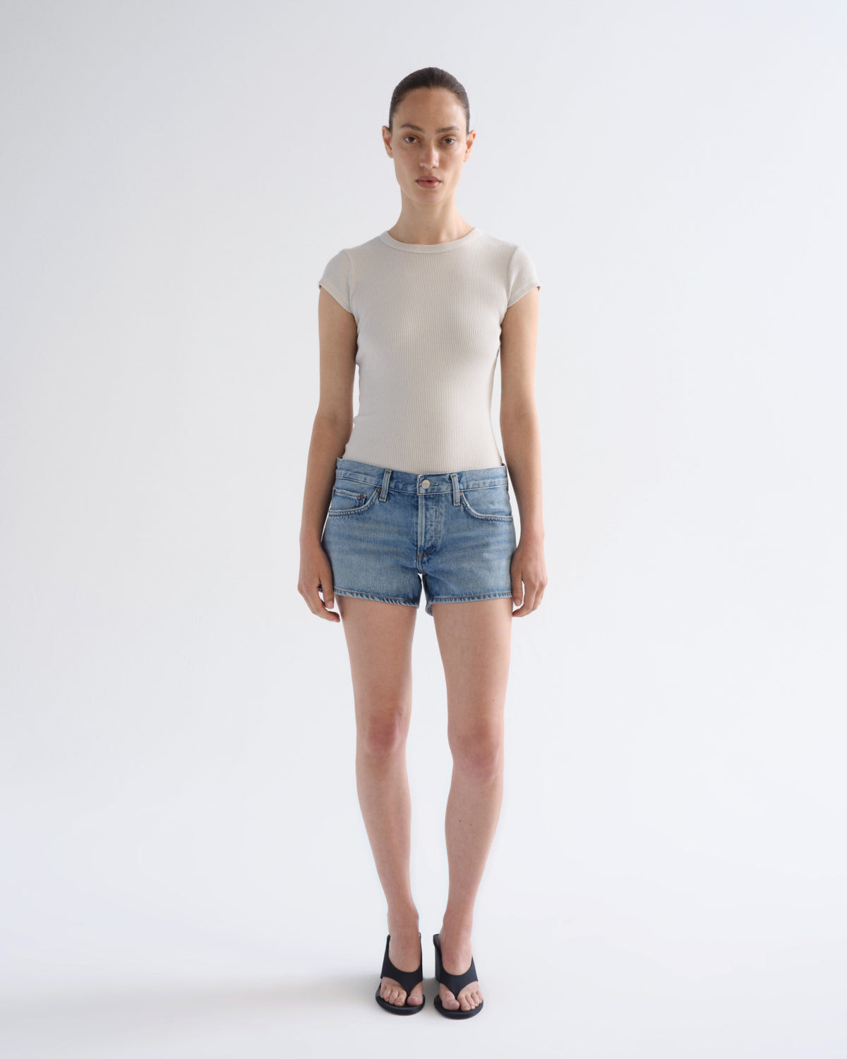 A model stands against a light gray studio background wearing a fitted short-sleeve beige top, blue denim shorts, and black sandals.