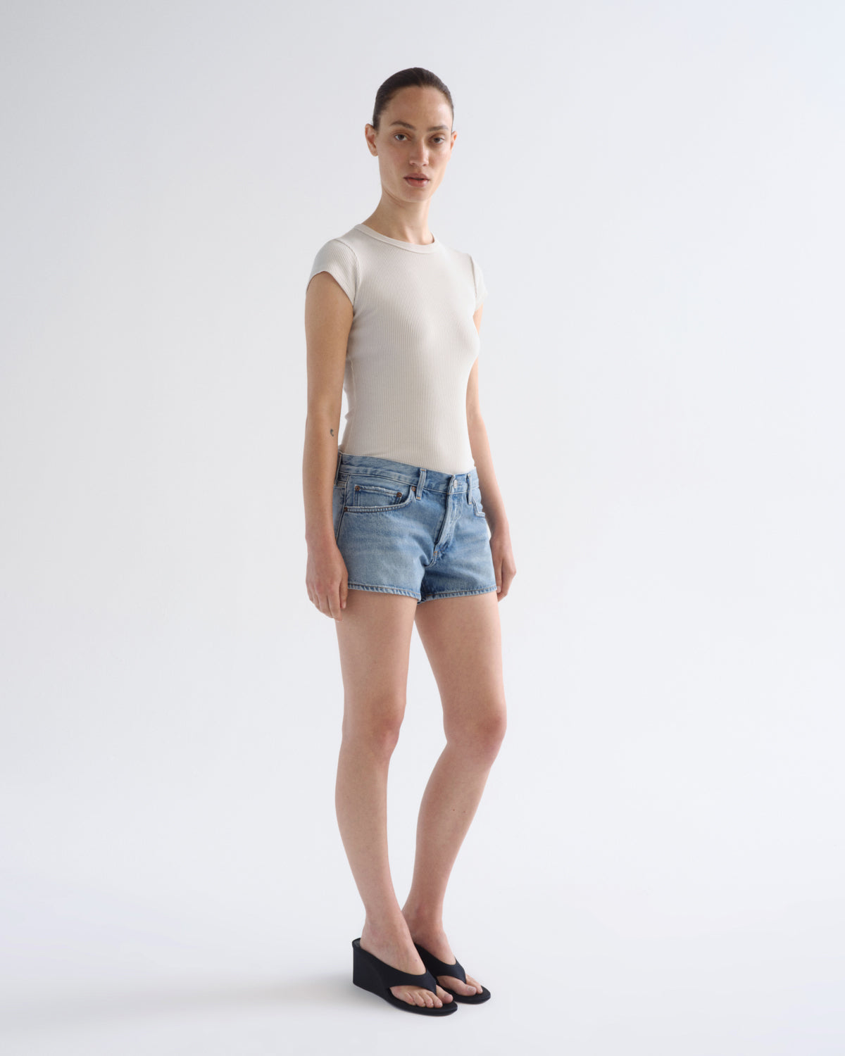 A model stands against a plain light gray studio background wearing a fitted short-sleeve cream top, light-wash denim shorts, and black wedge sandals.