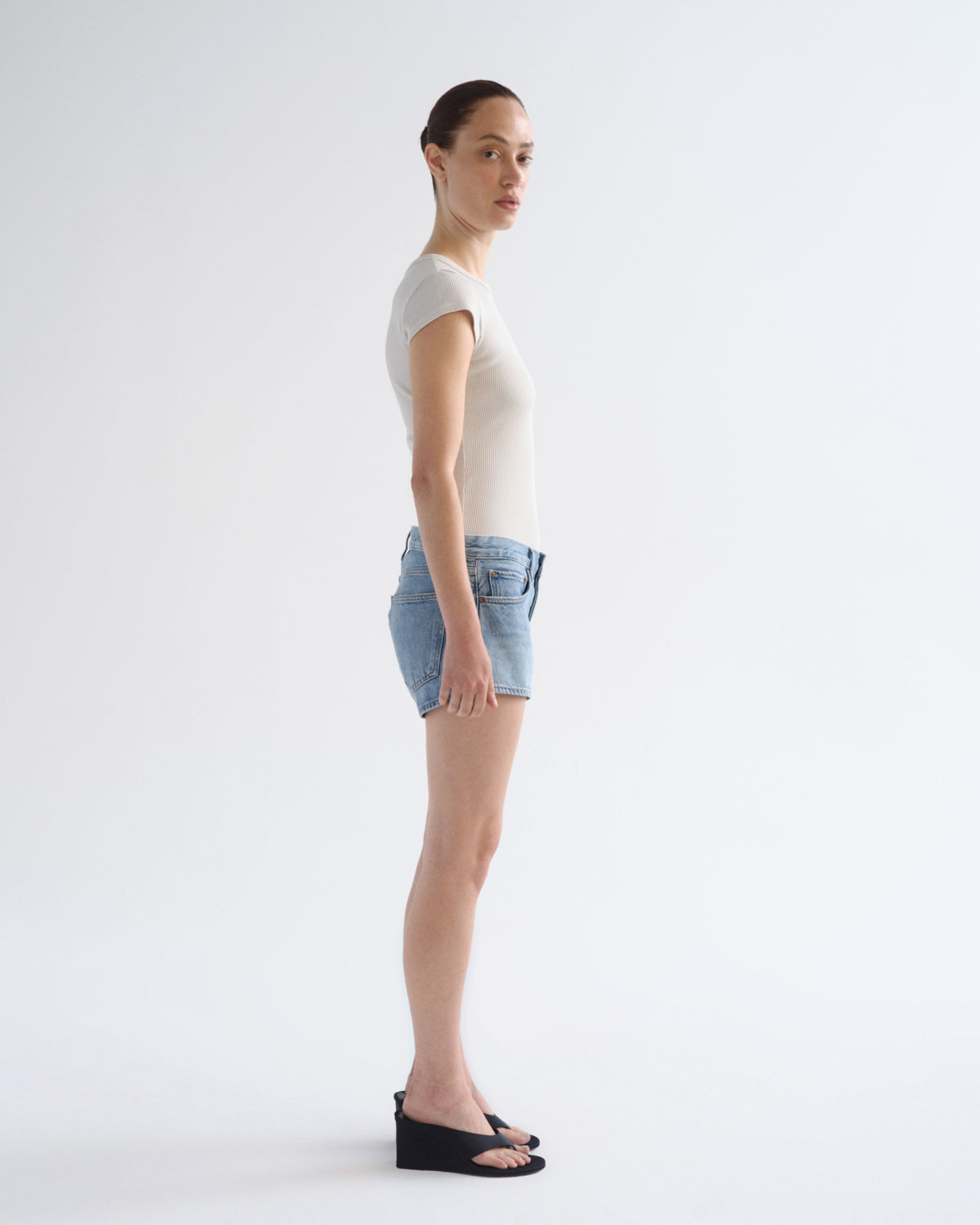 A woman stands in profile wearing a fitted white short-sleeve top, light blue denim shorts, and black wedge sandals against a plain white studio background.