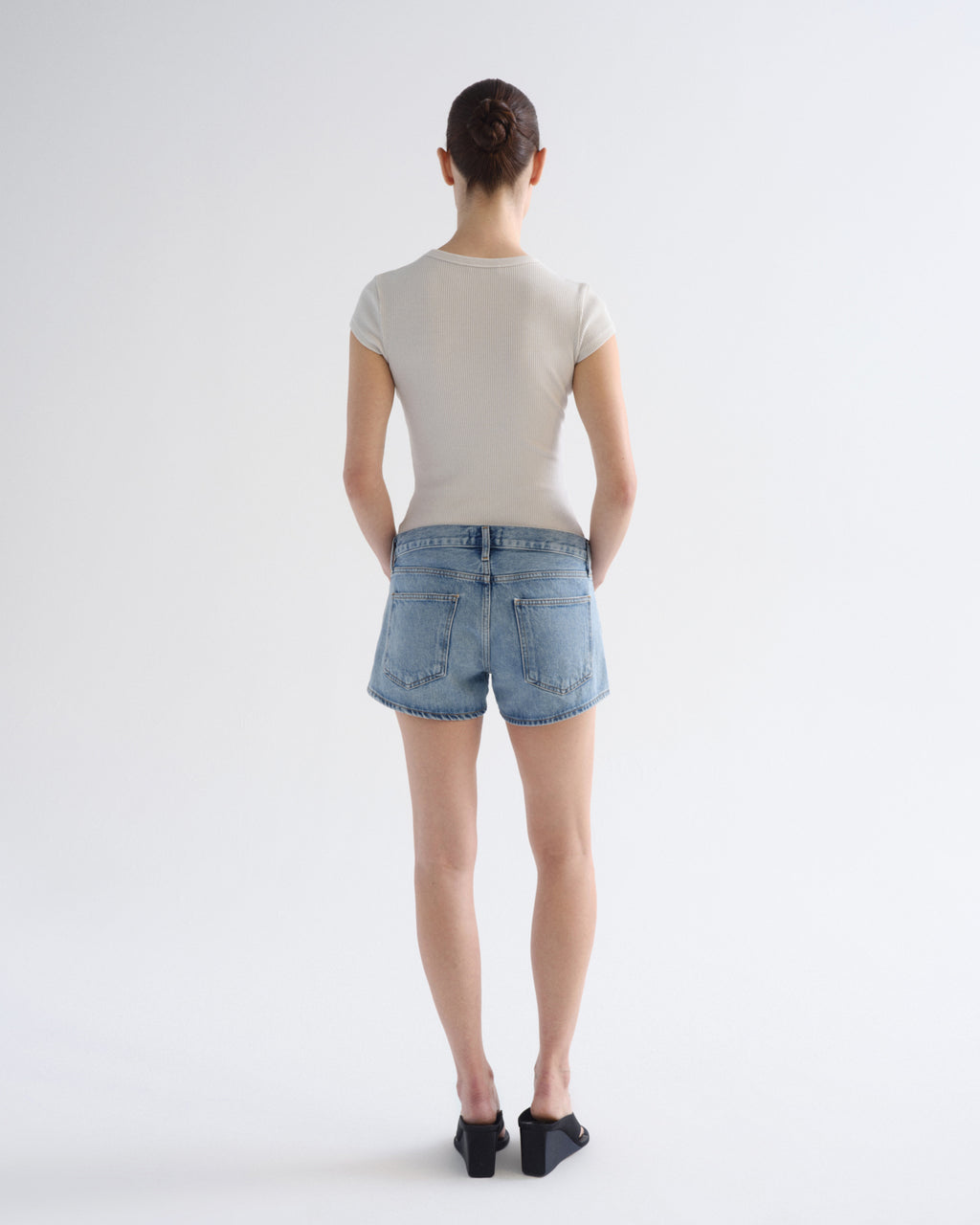 A woman is shown from behind wearing a light beige short-sleeve t-shirt, light-wash blue denim shorts, and black flat shoes against a plain white studio background.