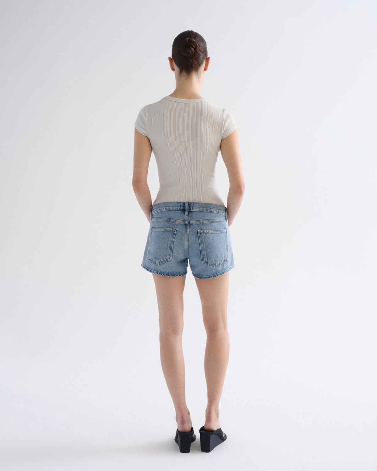 A woman is shown from behind wearing a light beige short-sleeve t-shirt, light-wash blue denim shorts, and black flat shoes against a plain white studio background.
