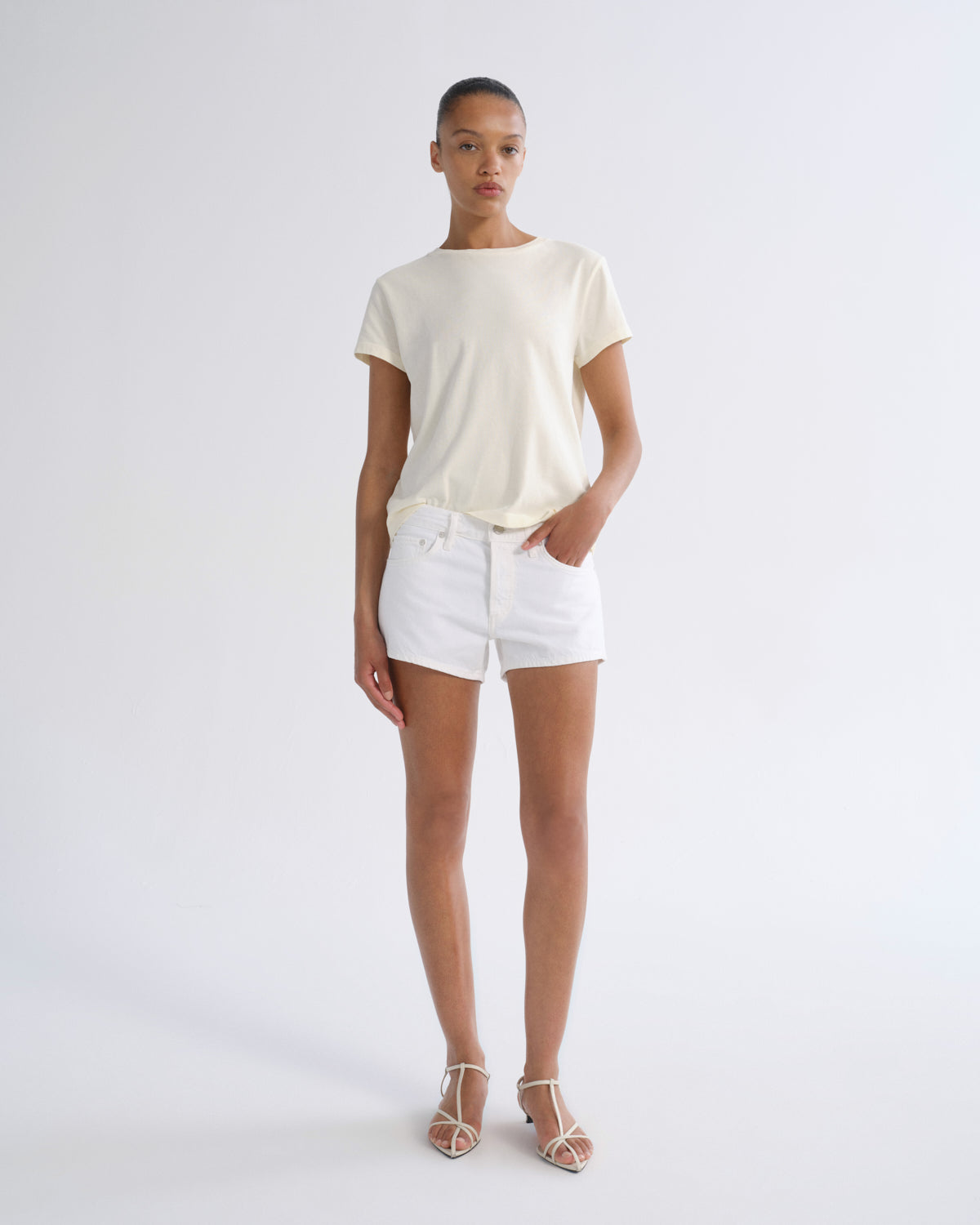 A model stands against a light gray studio background wearing a cream short-sleeve t-shirt, white denim shorts, and beige strappy high-heeled sandals.