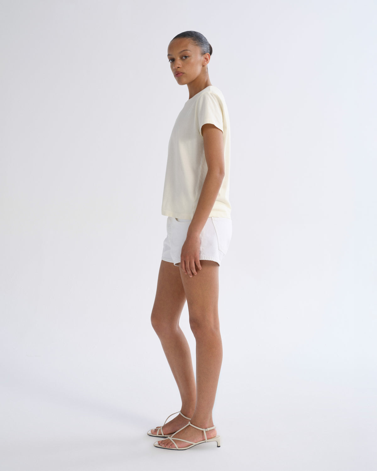 A model stands in profile wearing a cream short-sleeve t-shirt, white shorts, and strappy flat sandals against a plain white studio background.