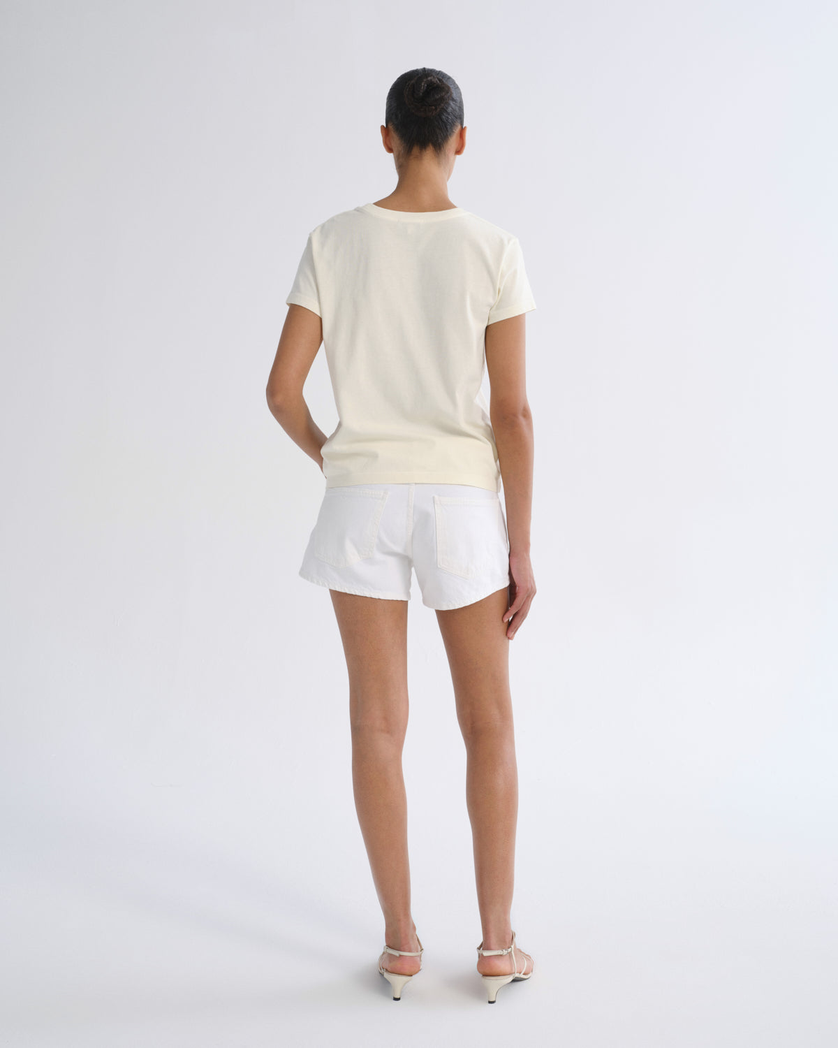 A woman viewed from the back wearing a cream short-sleeve t-shirt and white shorts, standing against a light gray studio background.