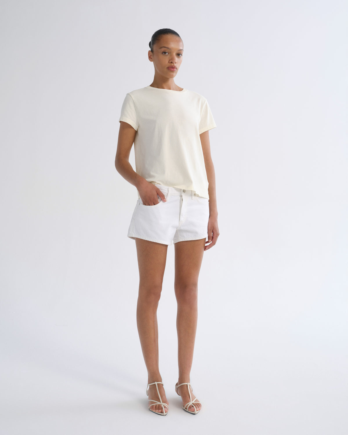 A model stands against a plain light gray studio background wearing a cream short-sleeve crewneck t-shirt, white shorts, and beige strappy high-heeled sandals.