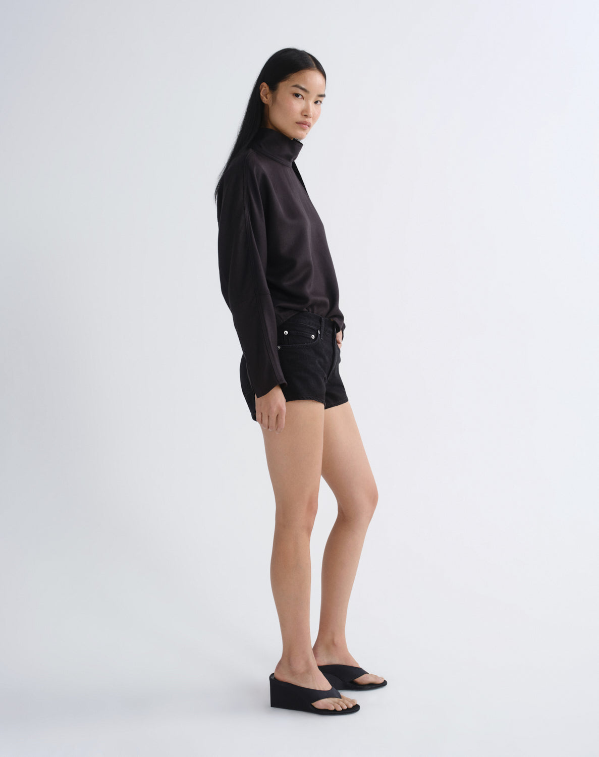A mode side view in a studio wearing a black long-sleeve mock-neck top with a draped collar detail, black shorts, and black wedge sandals against a light gray background.