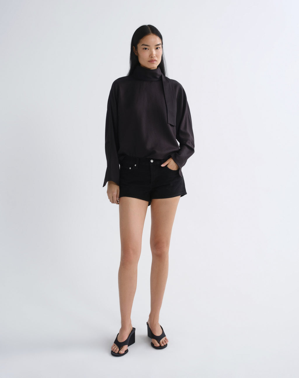 A model stands against a light gray studio background wearing a black long-sleeve high-neck blouse, black shorts, and black wedge sandals.