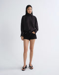 A model stands against a light gray studio background wearing a black long-sleeve high-neck blouse, black shorts, and black wedge sandals.