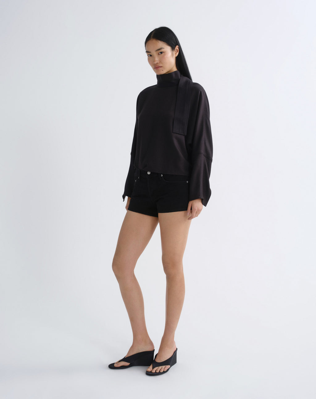 A model stands in a studio wearing a black long-sleeve mock-neck top with a draped collar detail, black shorts, and black wedge sandals against a light gray background.