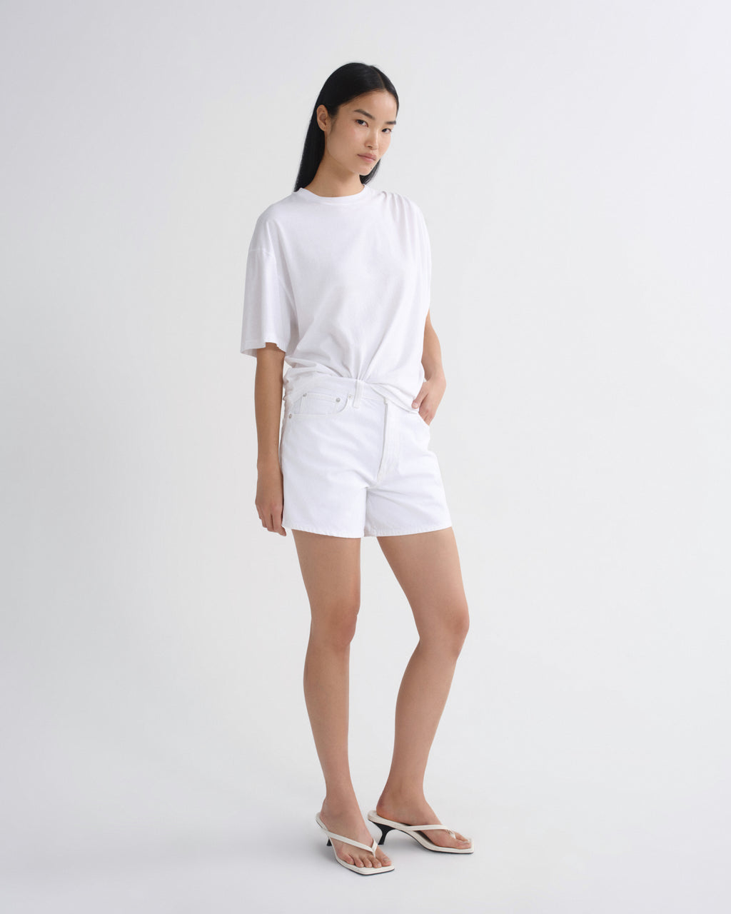 A woman modeling a white oversized short-sleeve t-shirt and matching white shorts, paired with white heeled sandals, against a plain light gray studio background.