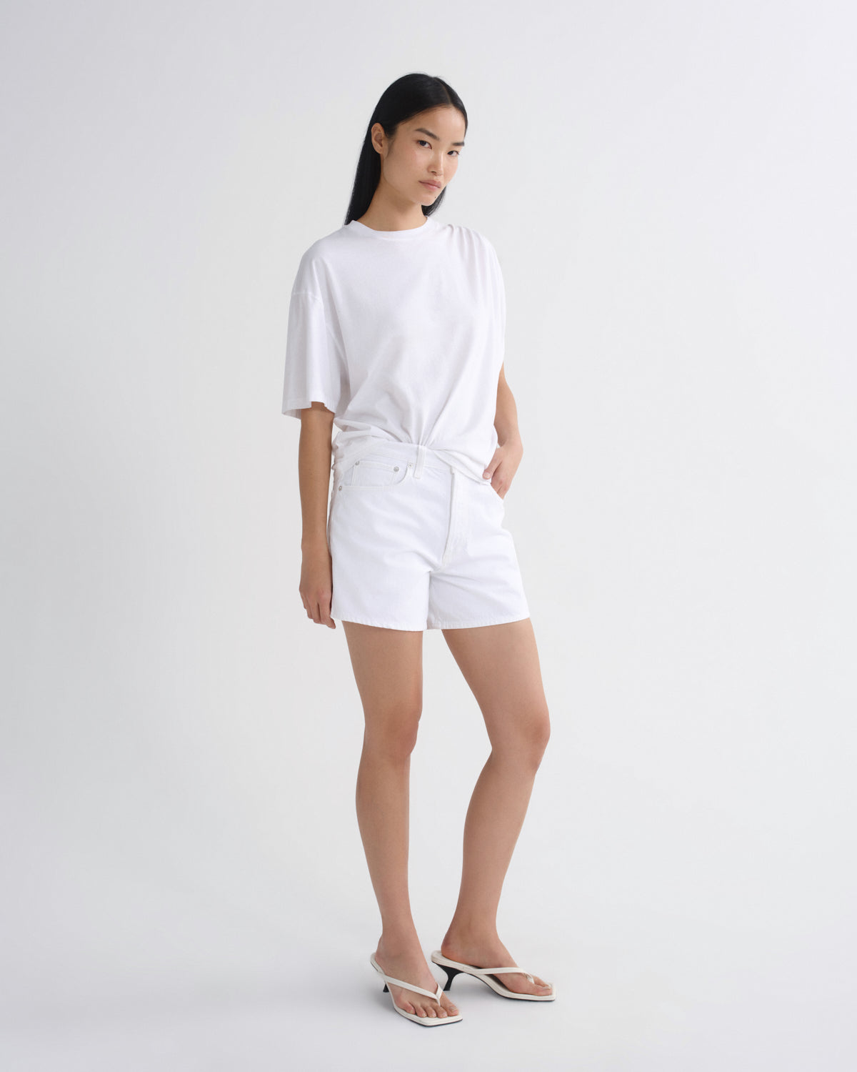 A woman modeling a white oversized short-sleeve t-shirt and matching white shorts, paired with white heeled sandals, against a plain light gray studio background.