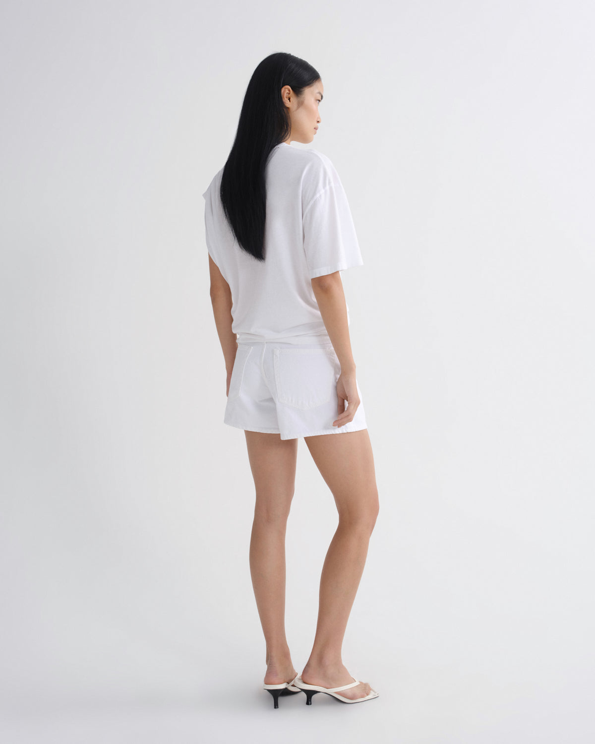 A woman with long black hair is shown from the back wearing a white oversized t-shirt and matching white shorts, paired with white heeled sandals, against a plain light gray studio background.