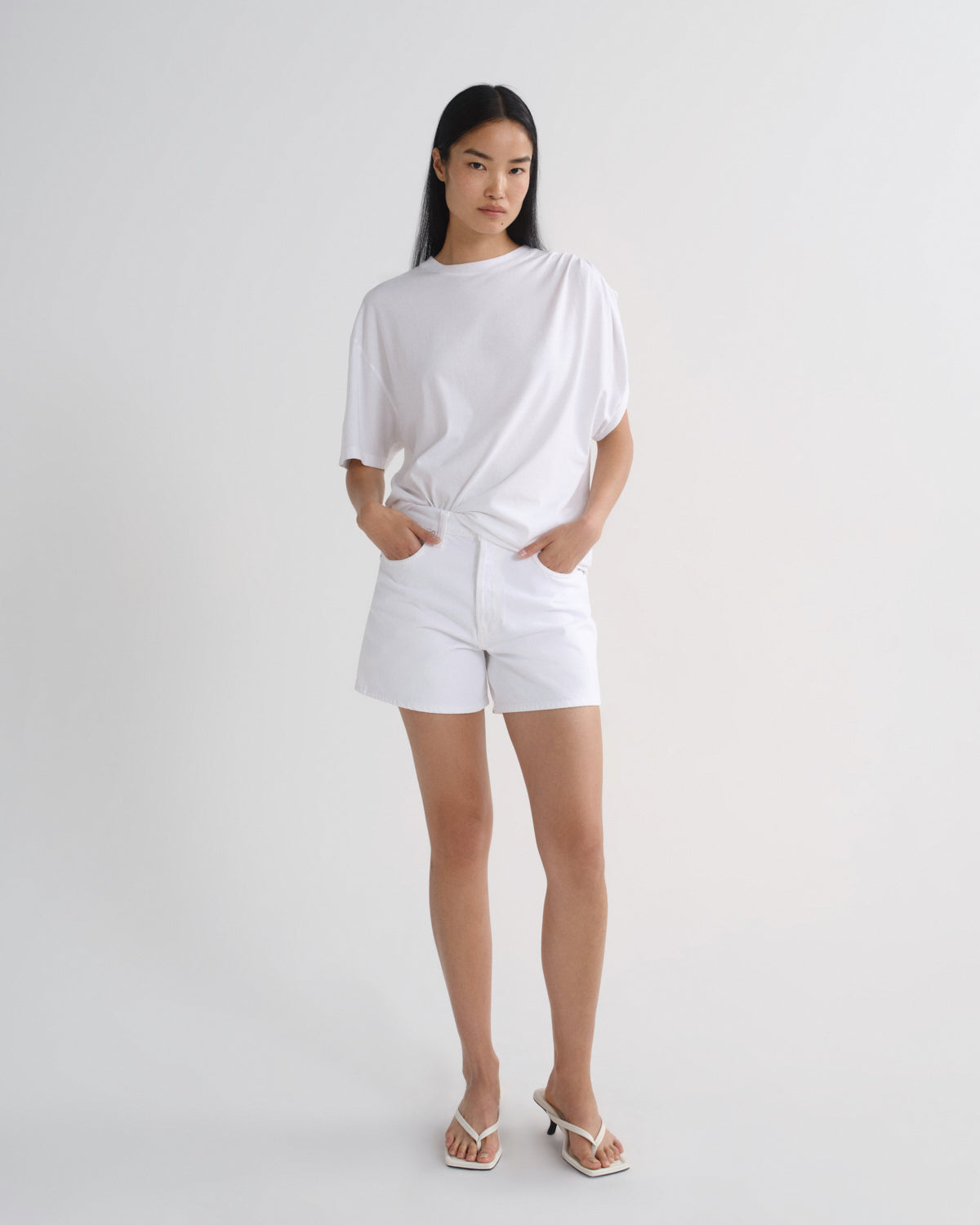 A woman modeling a white oversized short-sleeve t-shirt and matching white shorts against a plain light gray studio background.