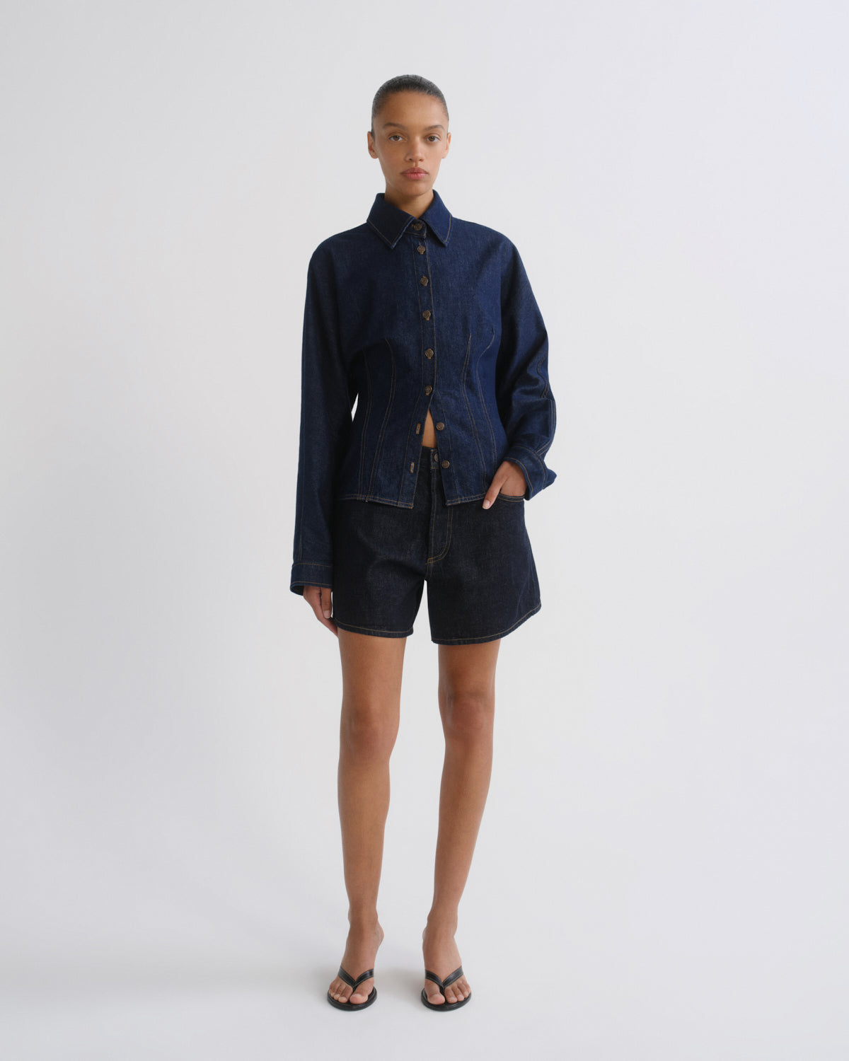 A model stands against a light gray studio background wearing a dark indigo denim button-up shirt and matching dark denim shorts, paired with black flip-flop sandals.