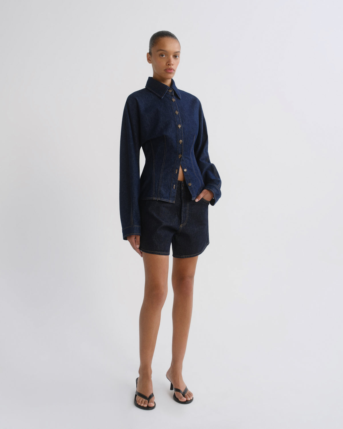 A model stands against a light gray studio background wearing a dark indigo denim long-sleeve button-up shirt and matching dark denim shorts, paired with black sandals.