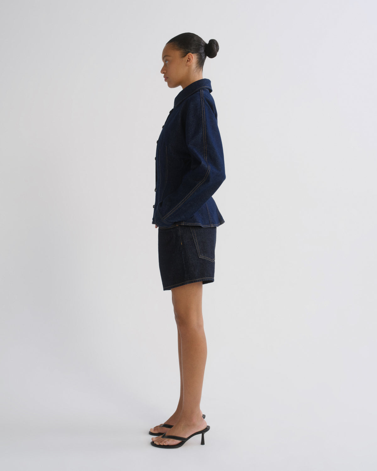 A model shown in profile wearing a dark indigo denim jacket and matching denim shorts, paired with black mule sandals, against a plain light gray studio background.