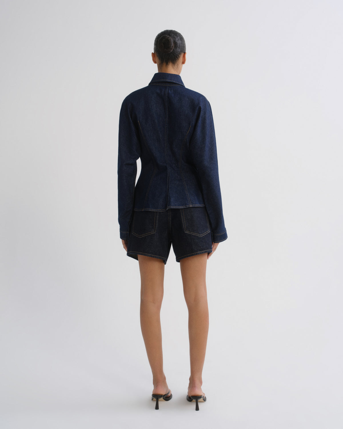 A model viewed from the back wearing a dark navy blue denim long-sleeve collared shirt and matching dark denim shorts, paired with black heeled sandals, against a plain light gray studio background.