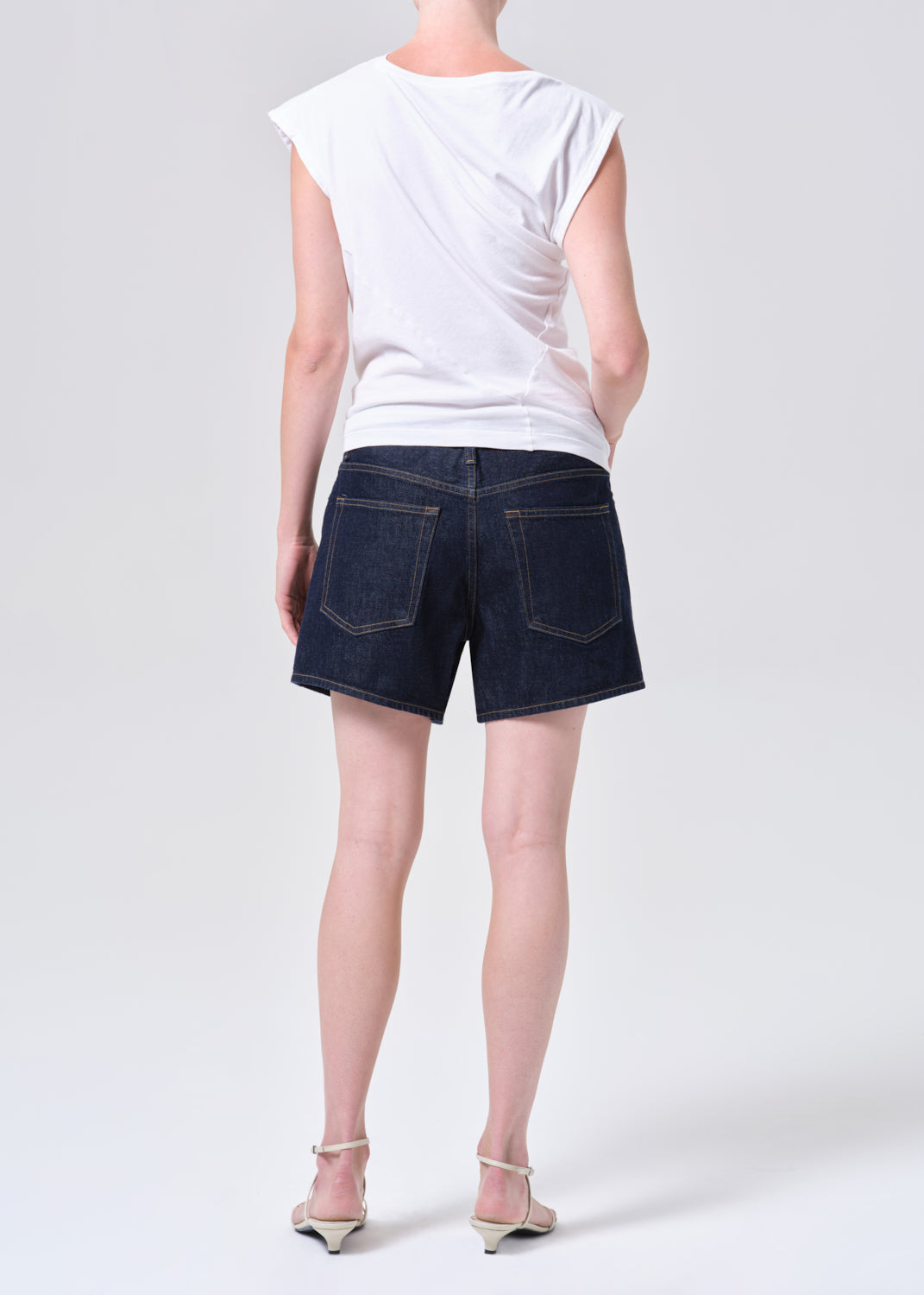 Person wearing a white sleeveless top and dark denim shorts on a plain background