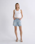 A woman modeling a white ribbed tank top and light-wash blue denim shorts against a plain light gray studio background.