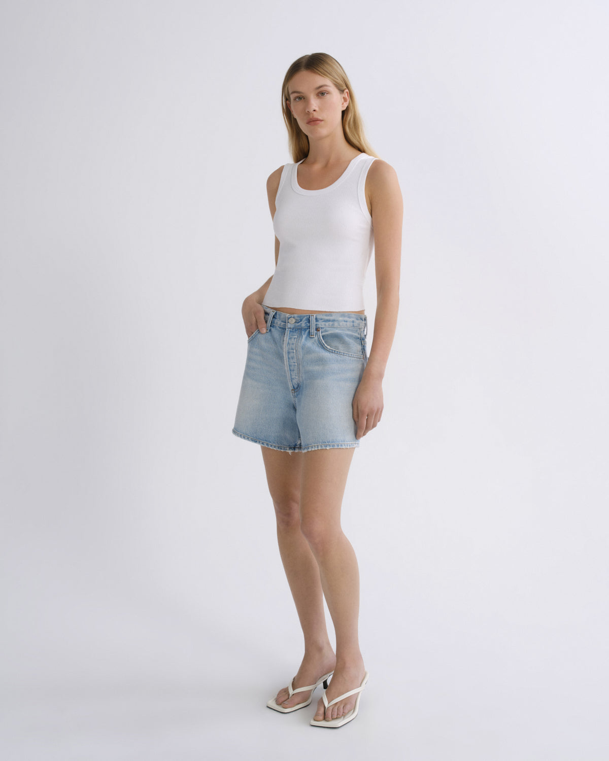 A woman modeling a white ribbed tank top and light-wash denim shorts against a clean, light gray studio background.