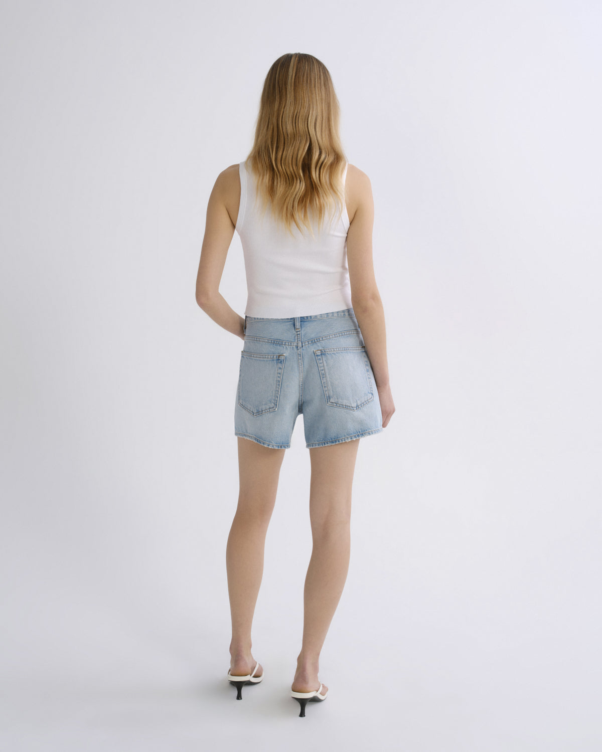 A woman is shown from behind wearing light-wash denim shorts, a white tank top, and white heeled sandals against a plain light gray studio background.