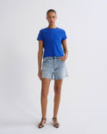 A model stands against a light gray studio background wearing a bright royal blue short-sleeve crewneck t-shirt, light-wash denim shorts, and black strappy sandals.