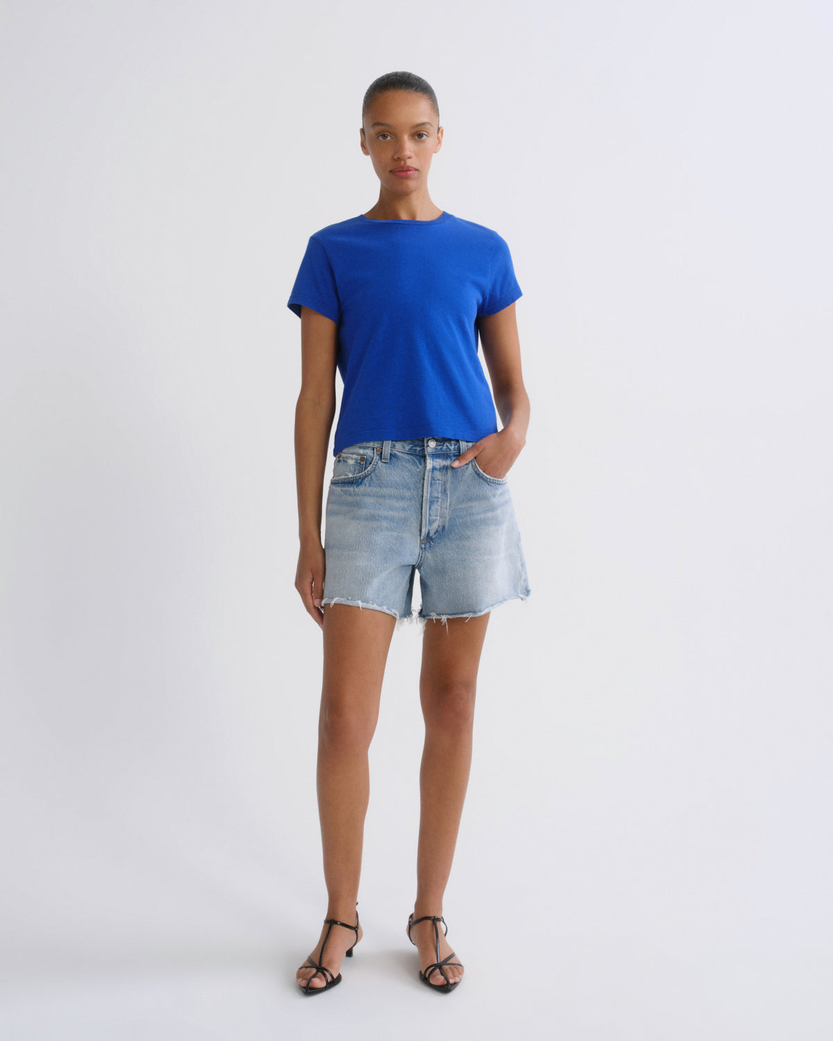 A model stands against a light gray studio background wearing a bright royal blue short-sleeve crewneck t-shirt, light-wash denim shorts, and black strappy sandals.