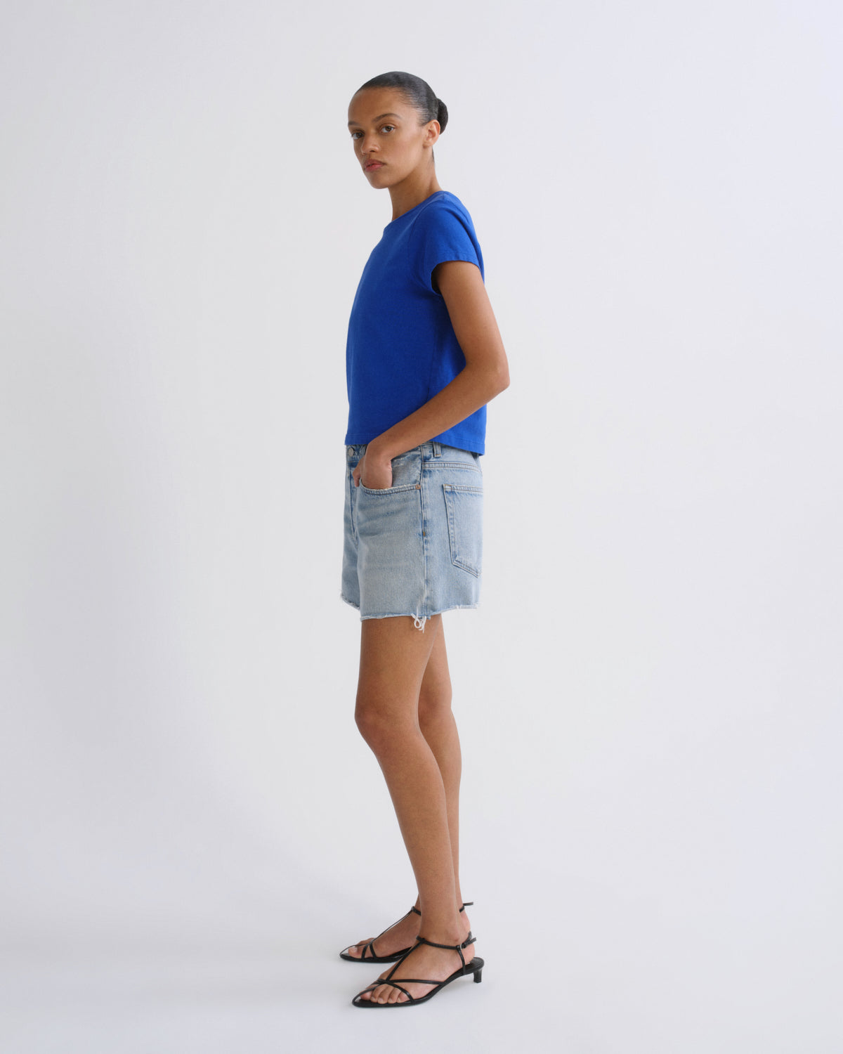 A model standing in profile wearing a bright blue short-sleeve t-shirt, a light-wash denim mini skirt, and black strappy sandals against a plain white studio background.