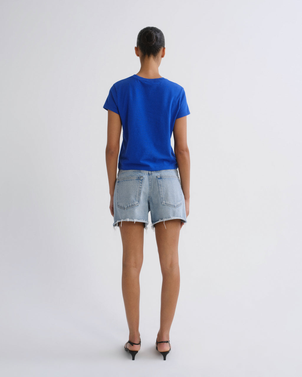 A woman viewed from behind wearing a bright blue short-sleeve t-shirt, light-wash denim shorts, and black pointed-toe heels against a plain light gray studio background.