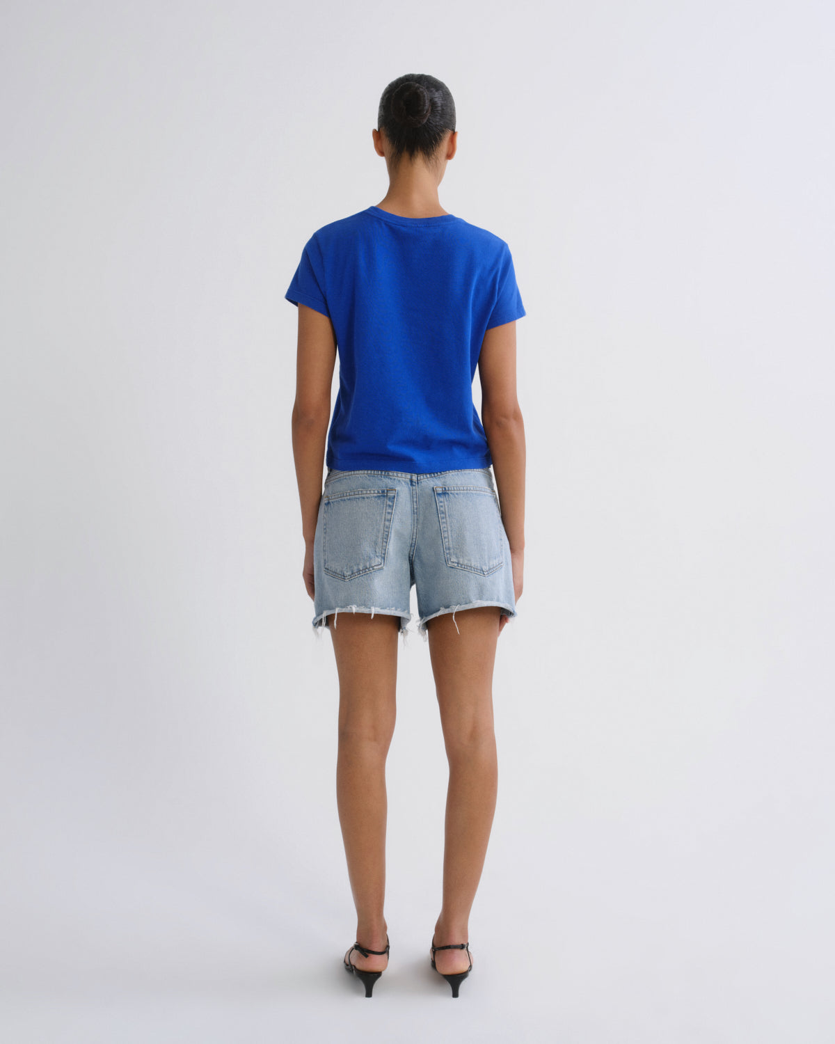 A woman viewed from behind wearing a bright blue short-sleeve t-shirt, light-wash denim shorts, and black pointed-toe heels against a plain light gray studio background.