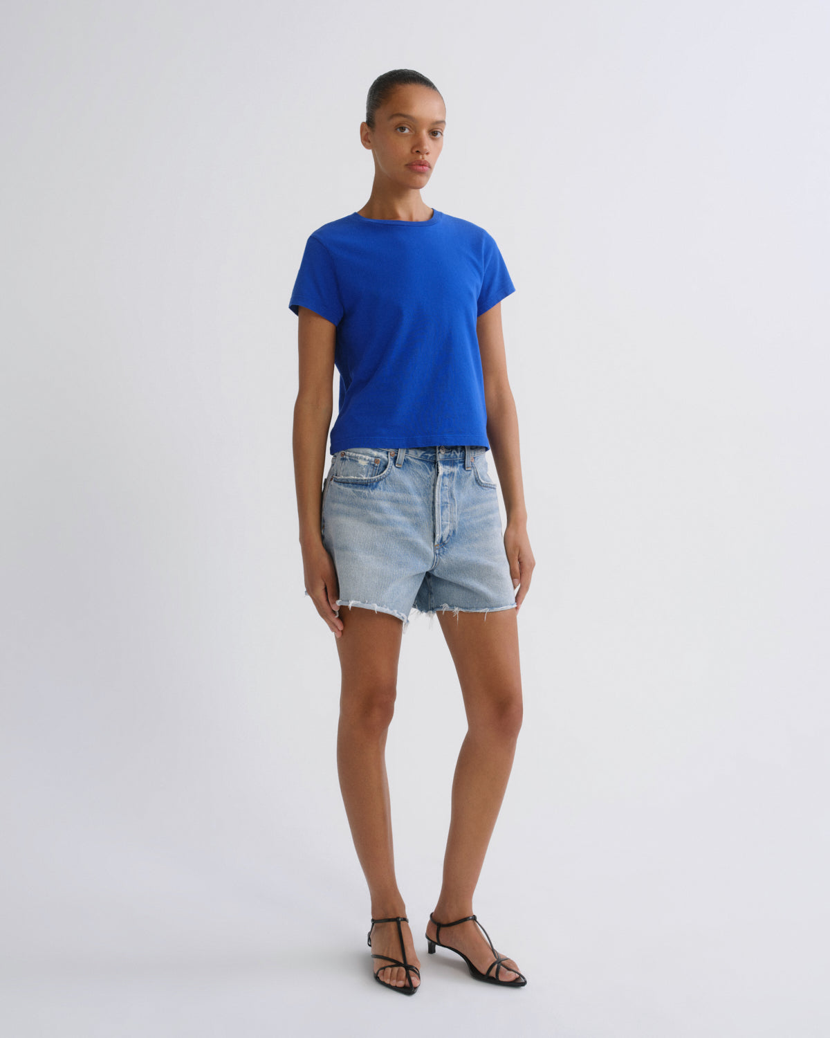 A model stands against a light gray studio background wearing a bright royal blue short-sleeve crewneck t-shirt, light-wash denim shorts, and black strappy sandals.
