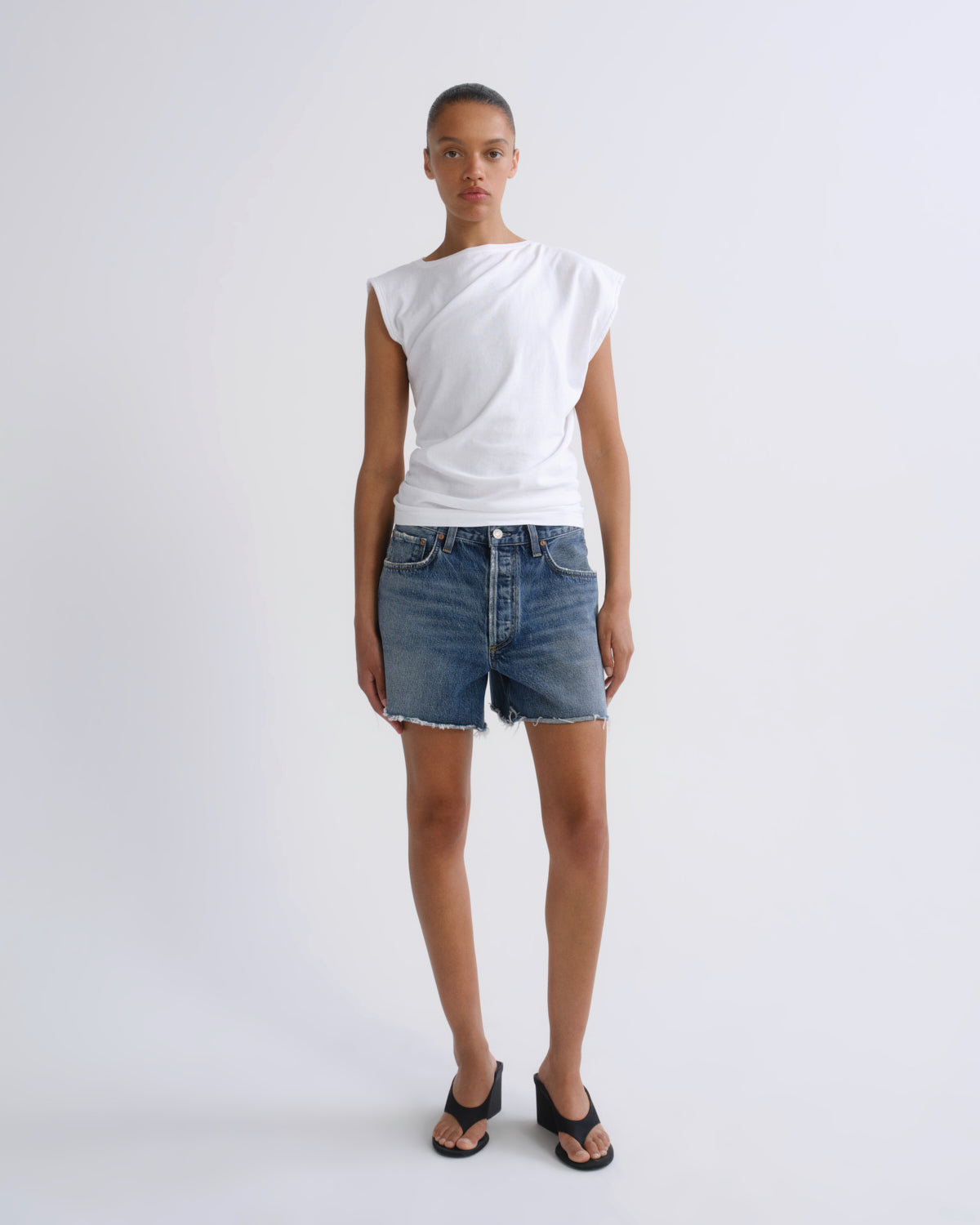 A model stands against a light gray studio background wearing a white asymmetrical sleeveless top, blue denim shorts, and black sandals.