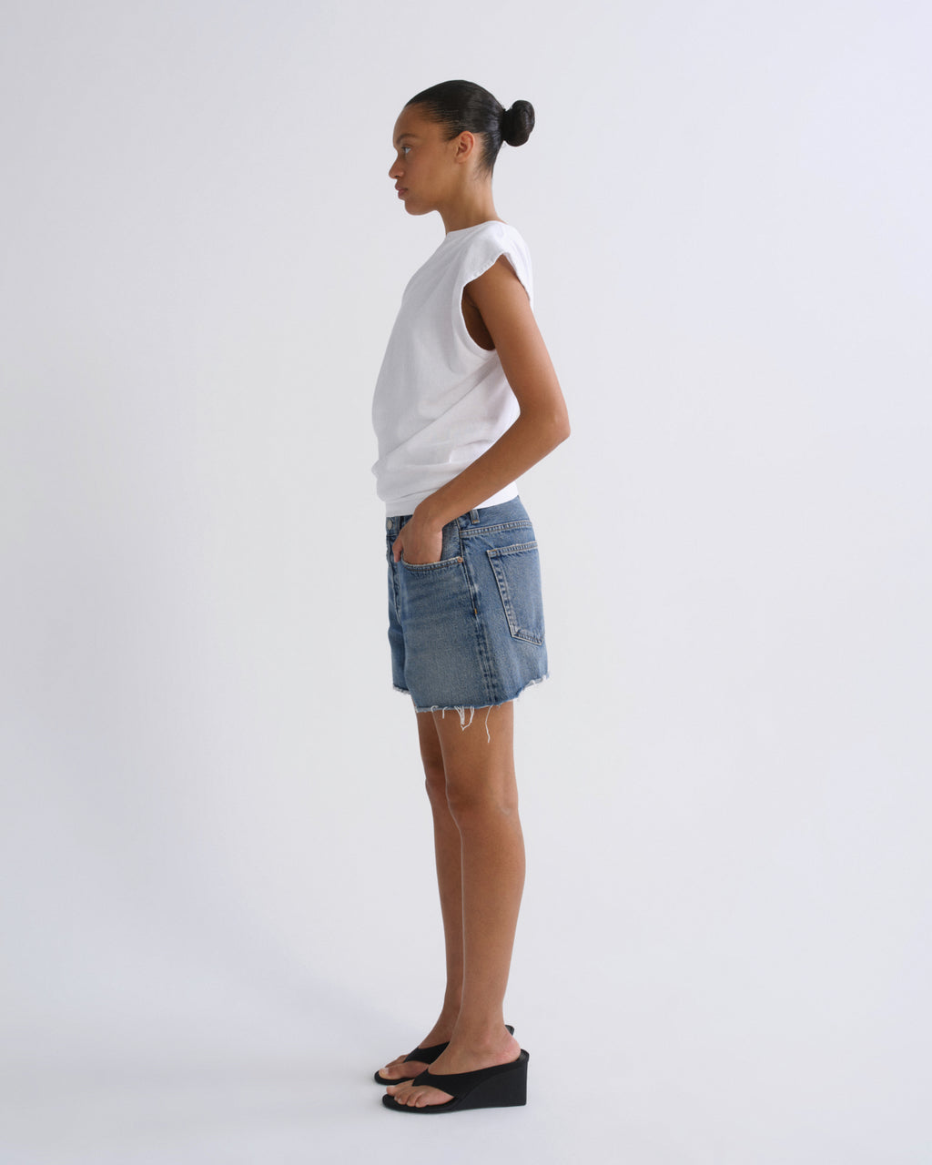 A model standing in profile wearing a white sleeveless top, a light-wash denim mini skirt, and black wedge sandals against a plain light gray studio background.
