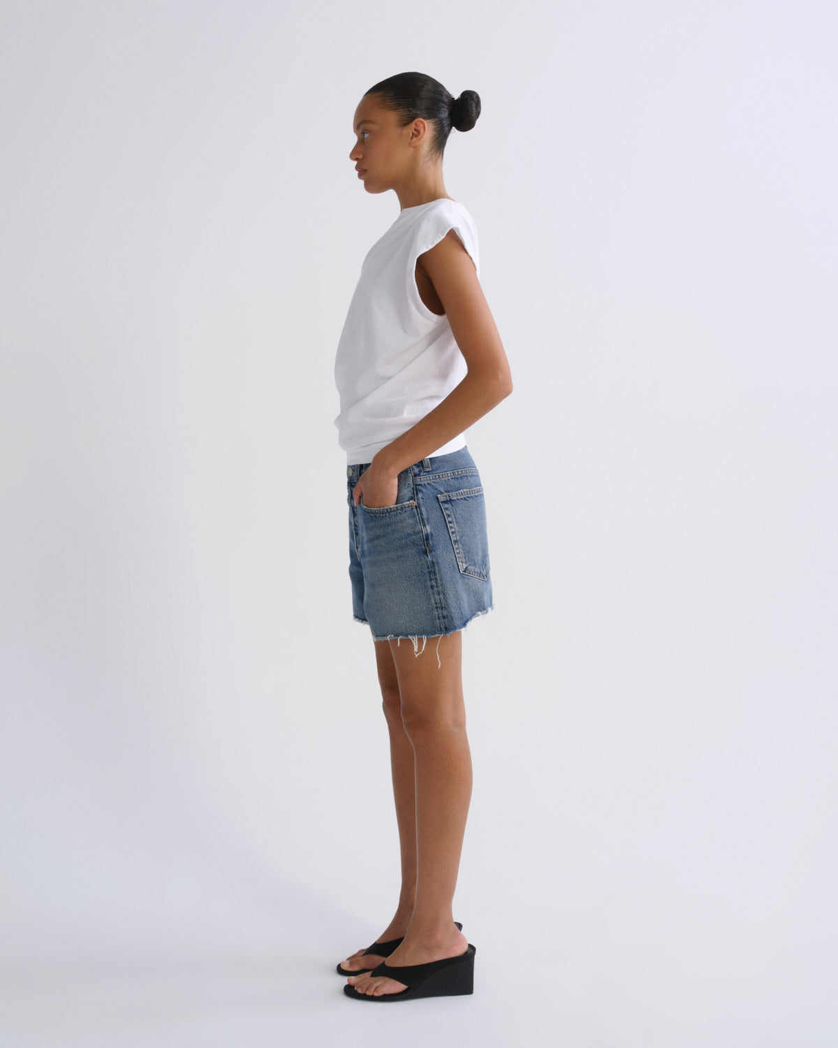 A model standing in profile wearing a white sleeveless top, a light-wash denim mini skirt, and black wedge sandals against a plain light gray studio background.