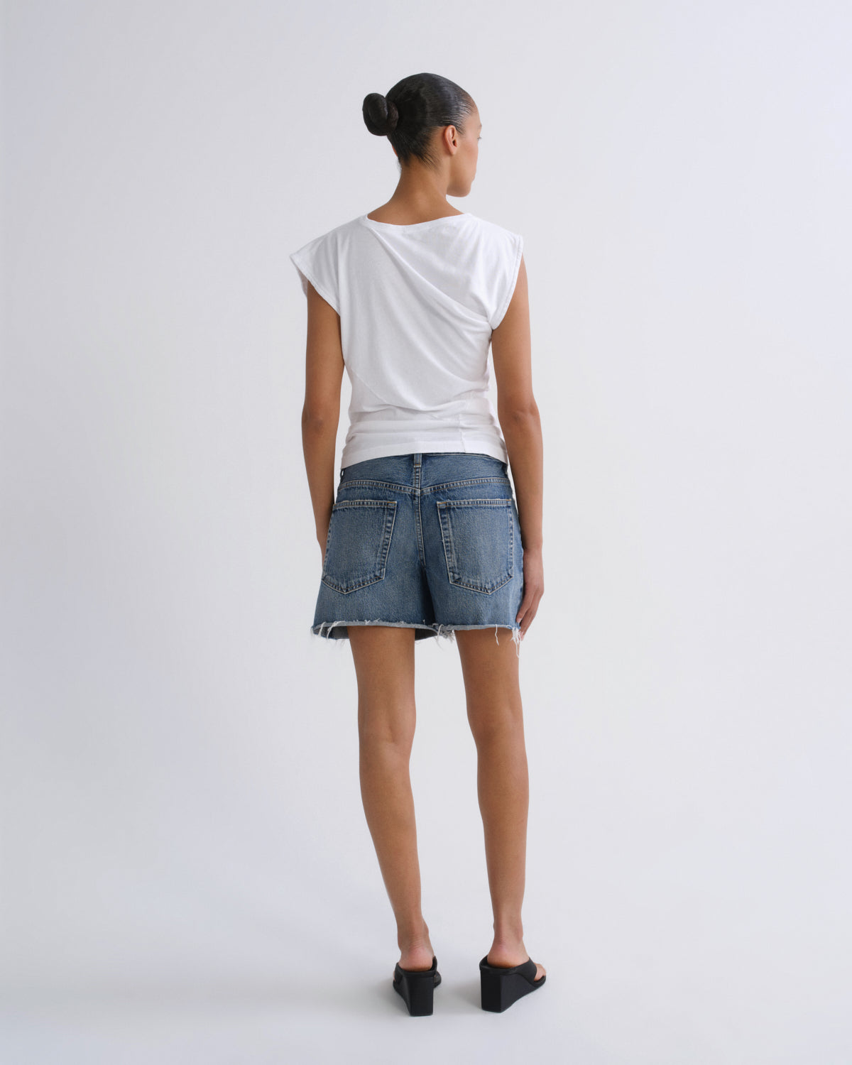 A model shown from the back wearing a white draped short-sleeve top, a blue denim mini skirt, and black wedge sandals against a light gray studio background.