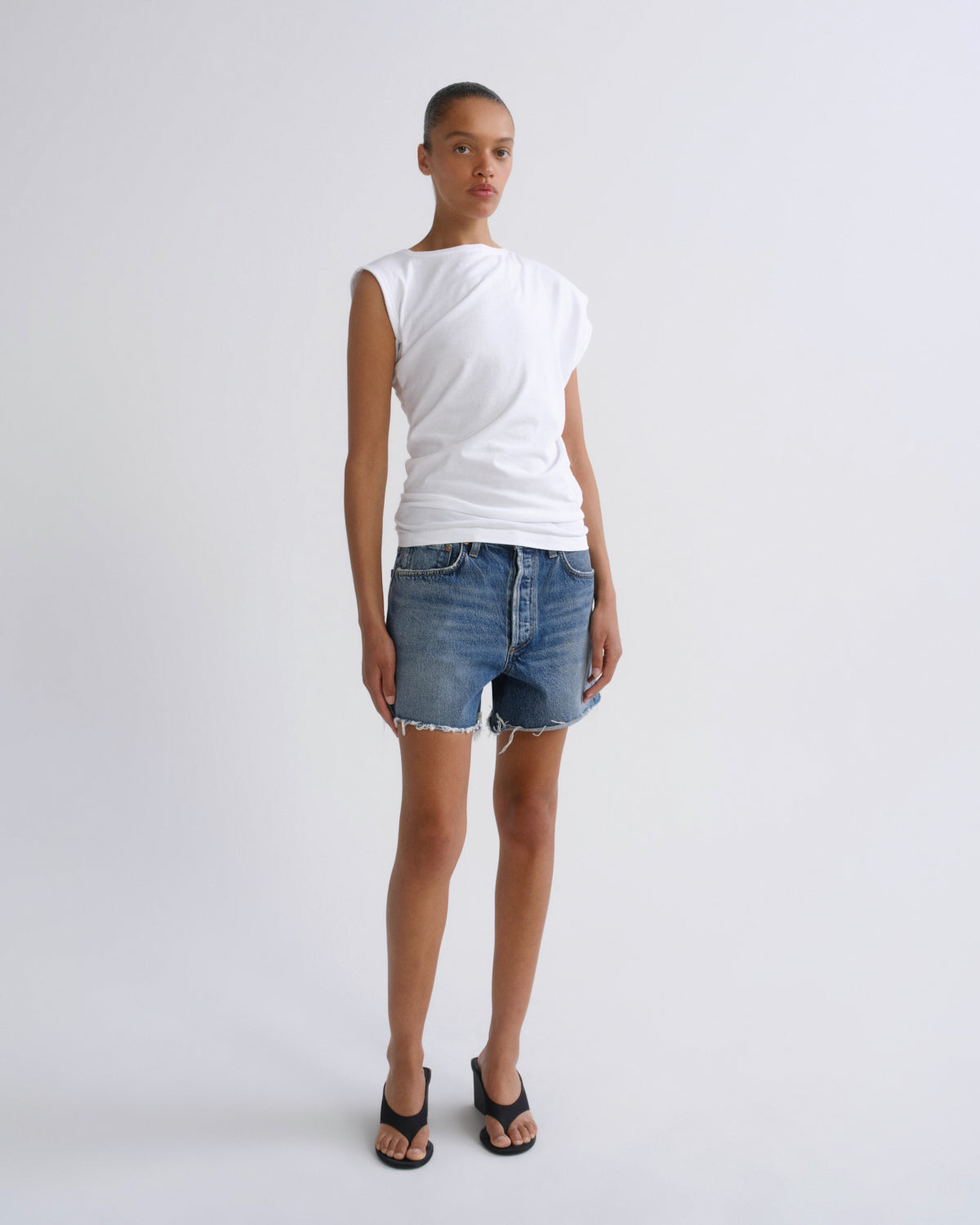 A model stands against a light gray studio background wearing a white sleeveless top, blue denim shorts, and black sandals.