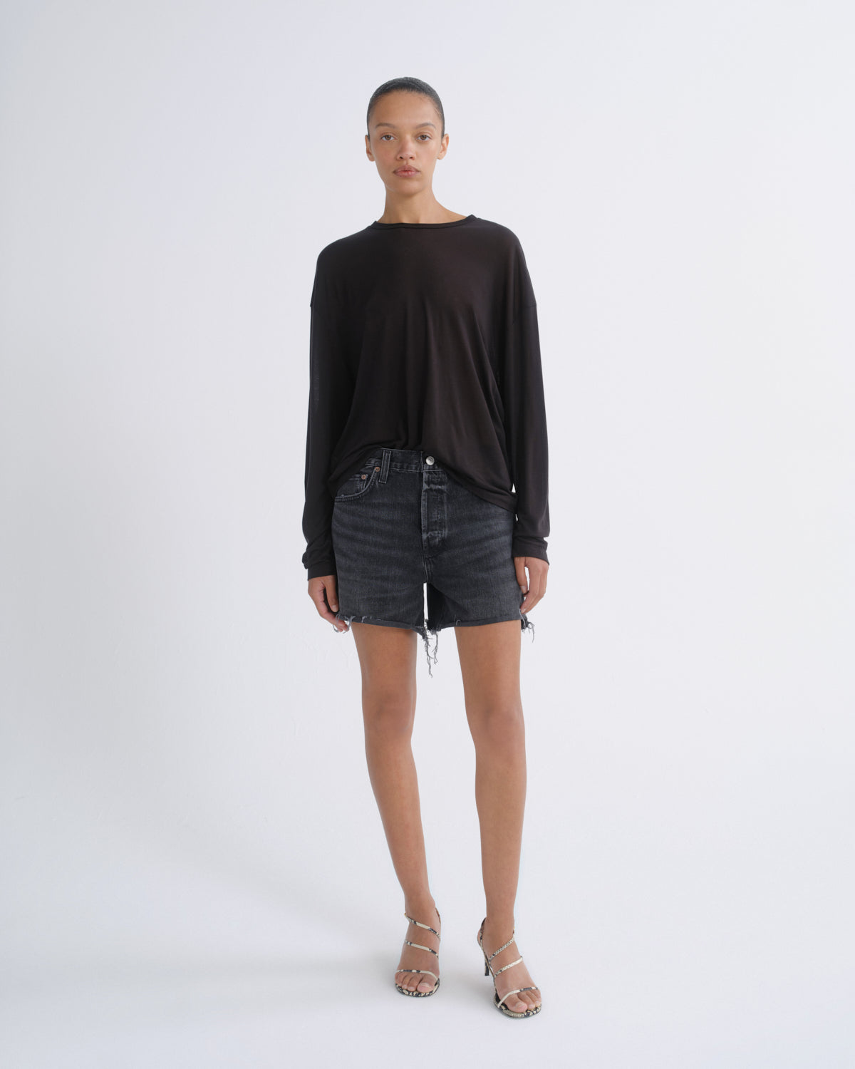 A model stands against a light gray studio background wearing a loose black long-sleeve top, dark gray denim shorts, and metallic strappy high-heeled sandals.