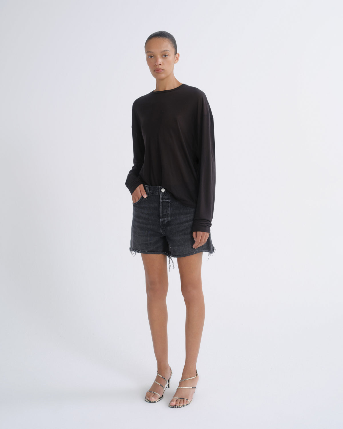 A model stands against a light gray studio background wearing a black long-sleeve knit top, dark gray frayed denim shorts, and silver strappy high-heeled sandals.
