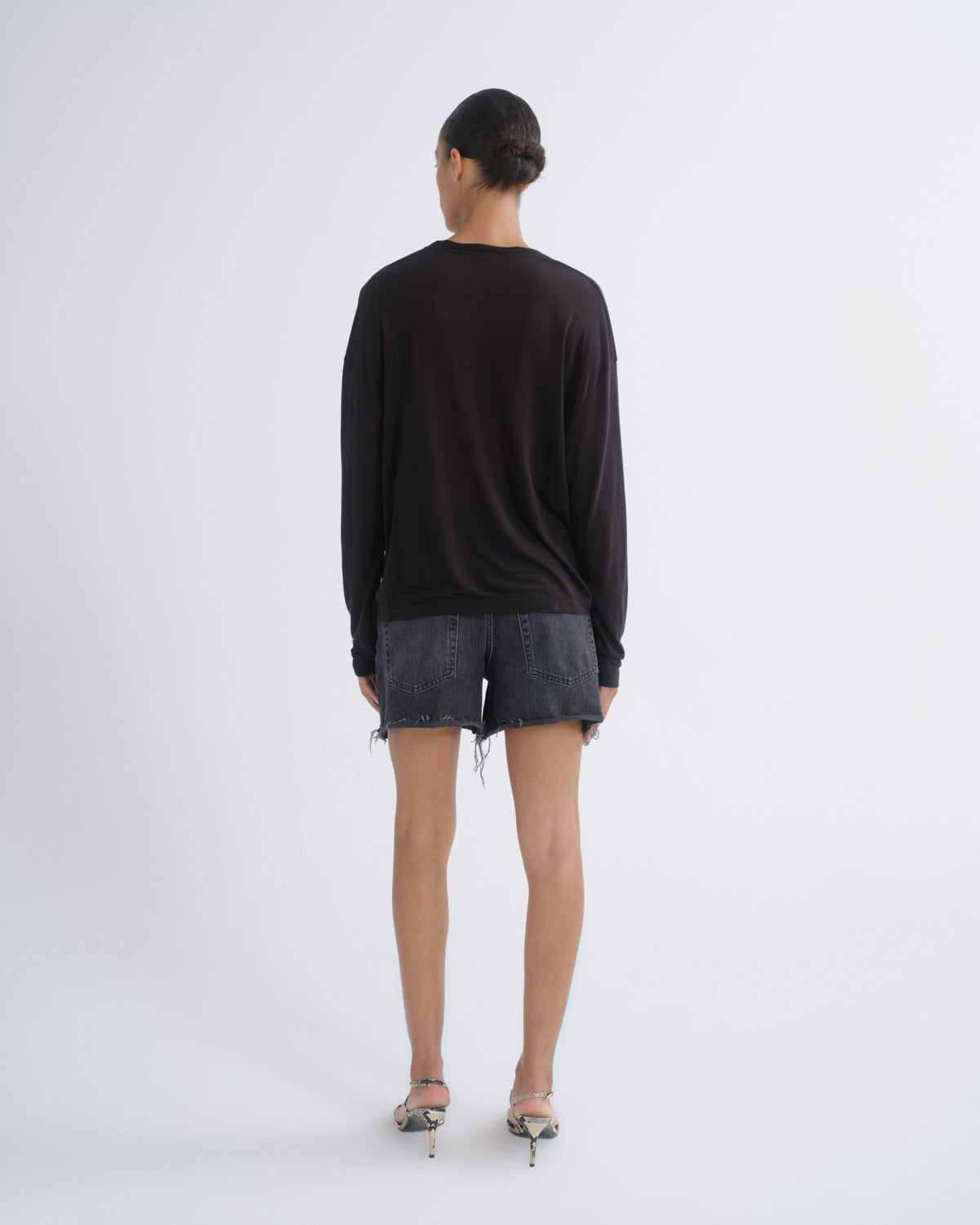 A model viewed from behind wearing a black long-sleeve top, dark gray denim shorts, and metallic high heels against a plain white studio background.