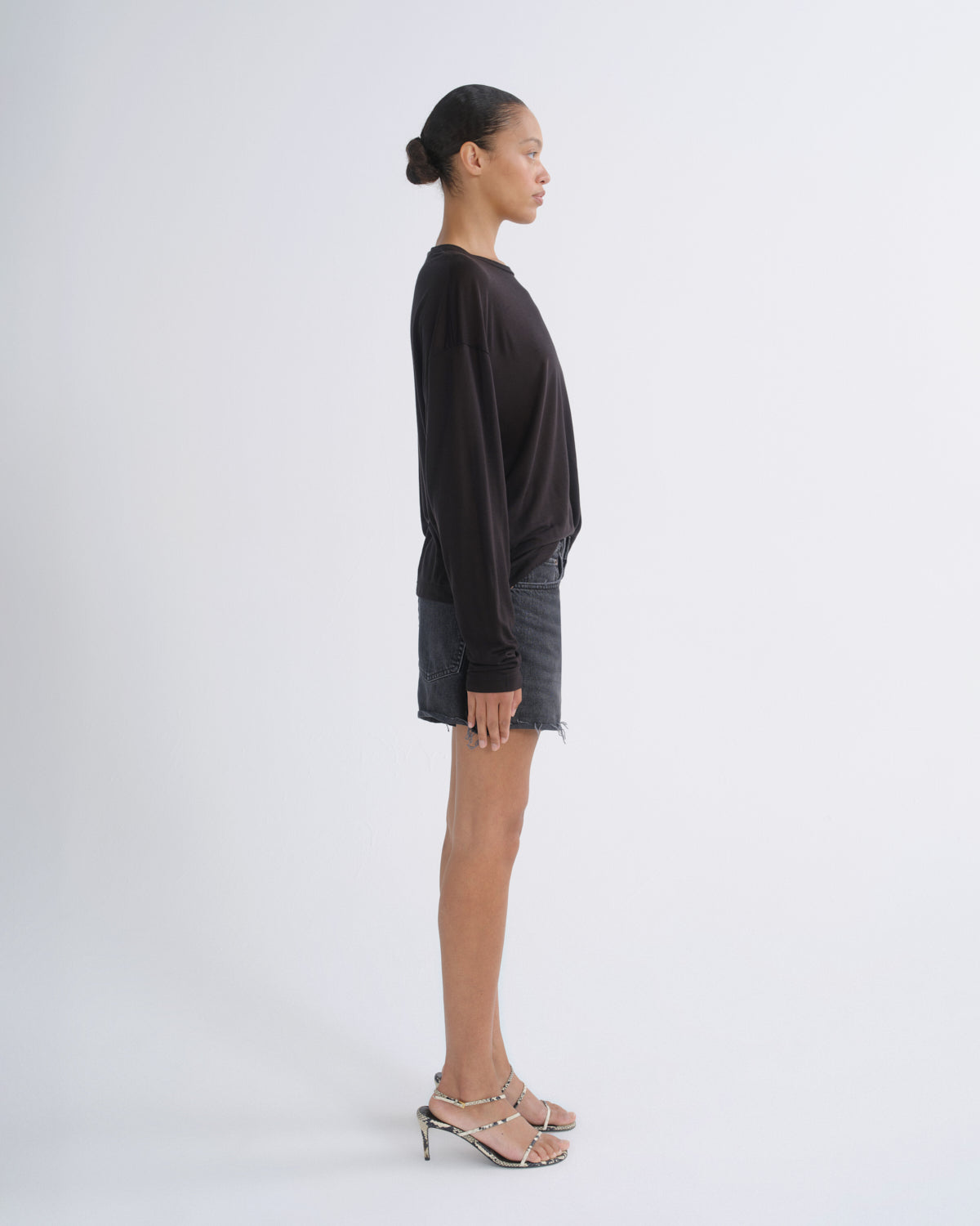 A model shown in profile wearing a black long-sleeve top, dark gray denim shorts, and metallic silver high-heeled sandals against a plain light gray studio background.