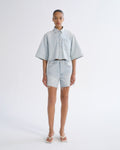 Woman wearing light blue denim shorts with a light blue short sleeve button-up shirt on a grey background.